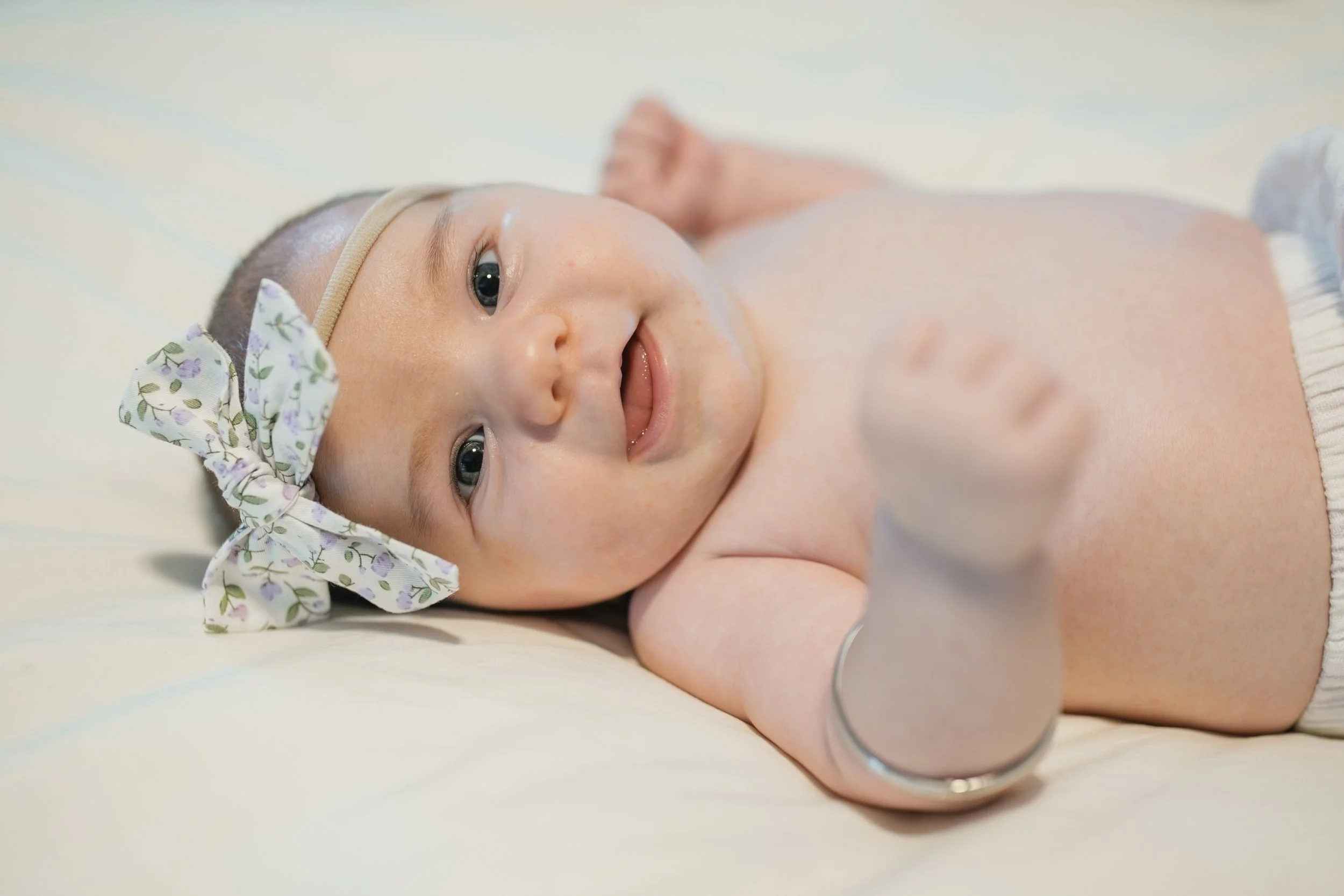 Close-up of a smiling baby lying down, wearing a floral headband, with a medical bracelet on their wrist.