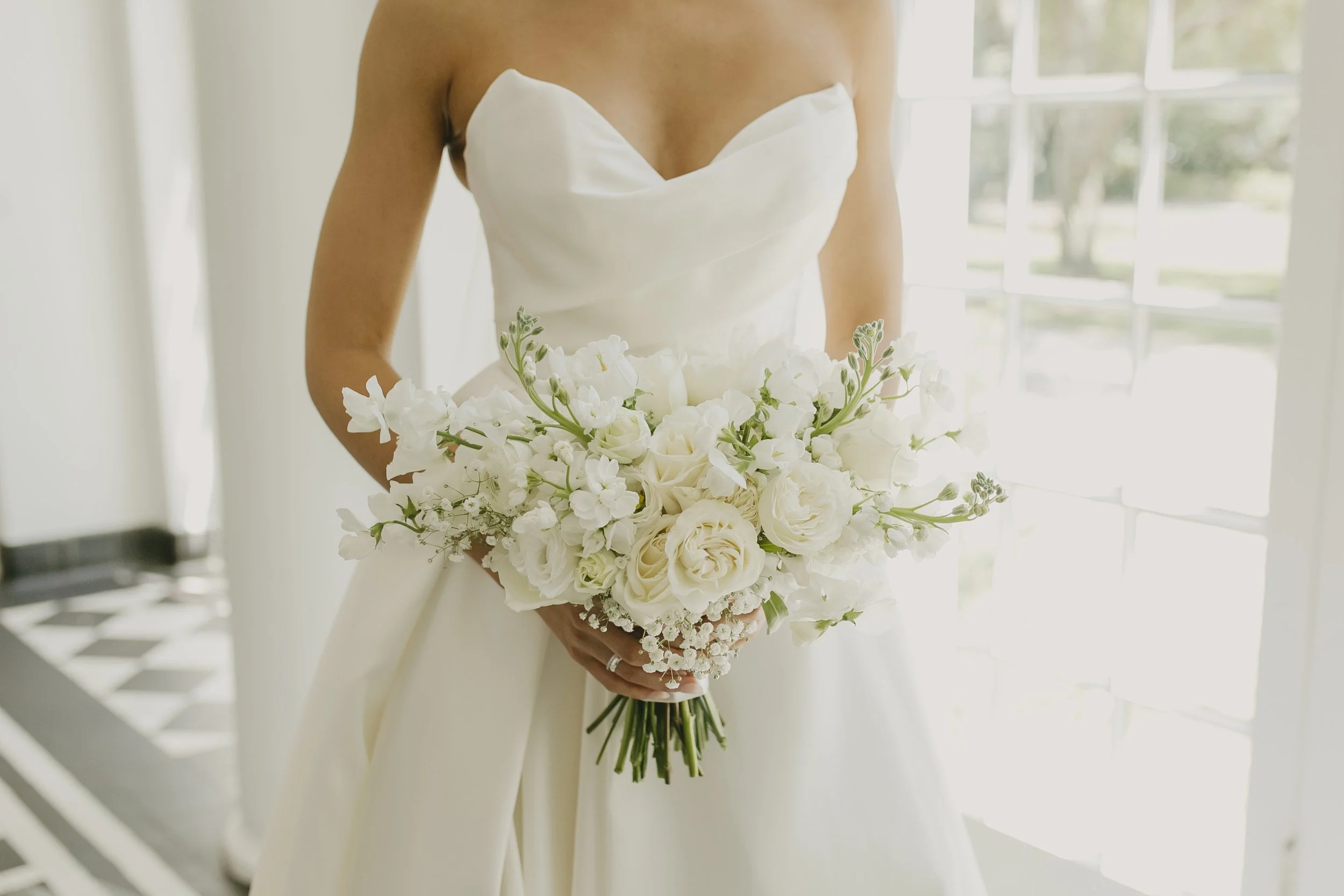 Bride wearing a white strapless wedding gown, holding a large bouquet of white flowers, standing indoors near a window.