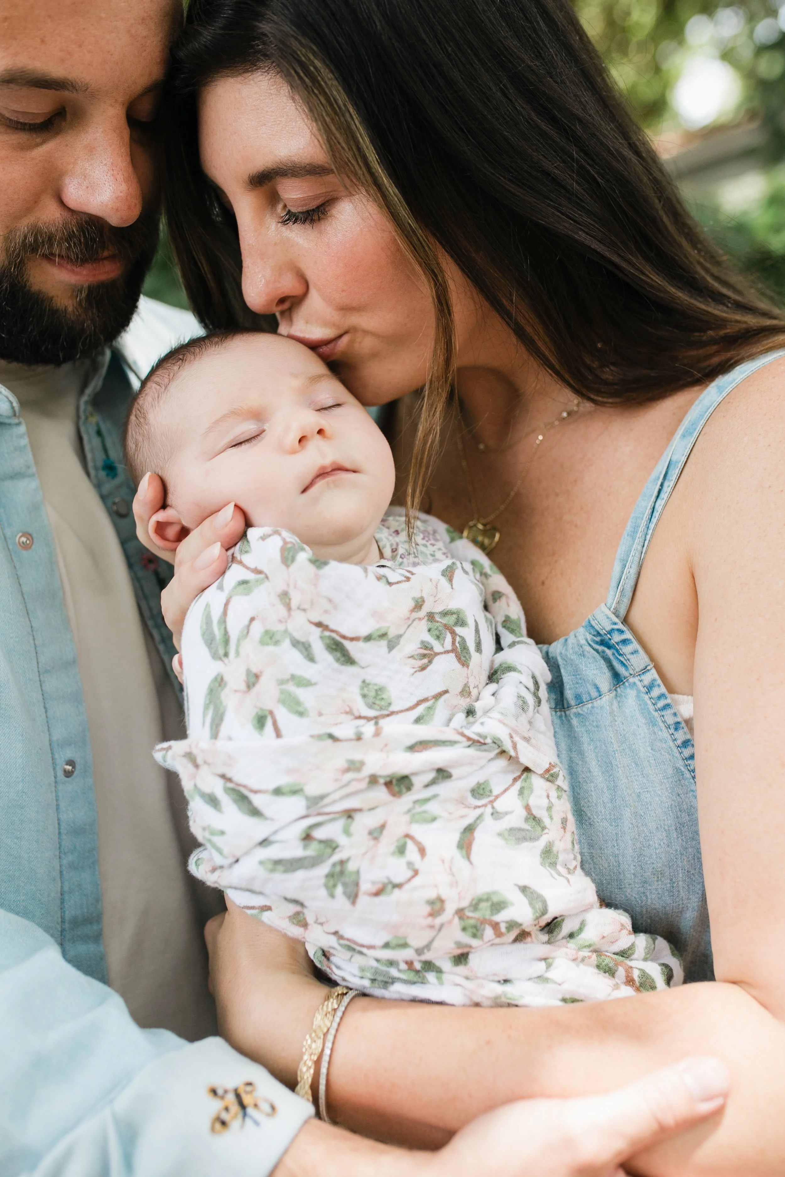 A family photo with a man, woman, and baby, all with their eyes closed, with the woman kissing the baby's forehead.