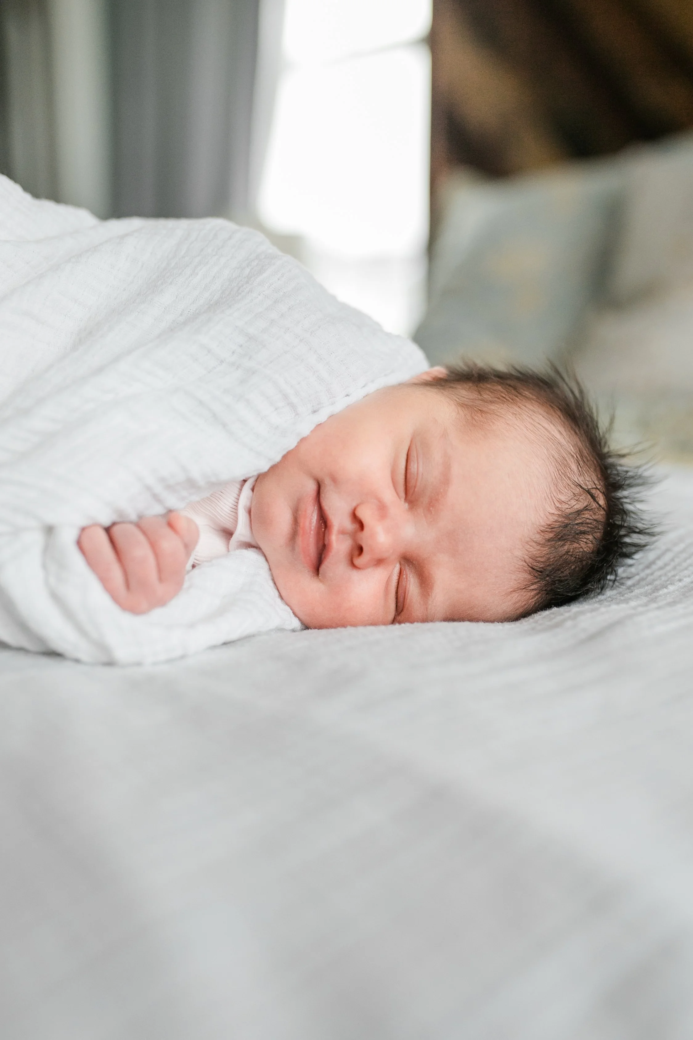 A sleeping baby with dark hair, wrapped in a white blanket, resting on a soft surface.
