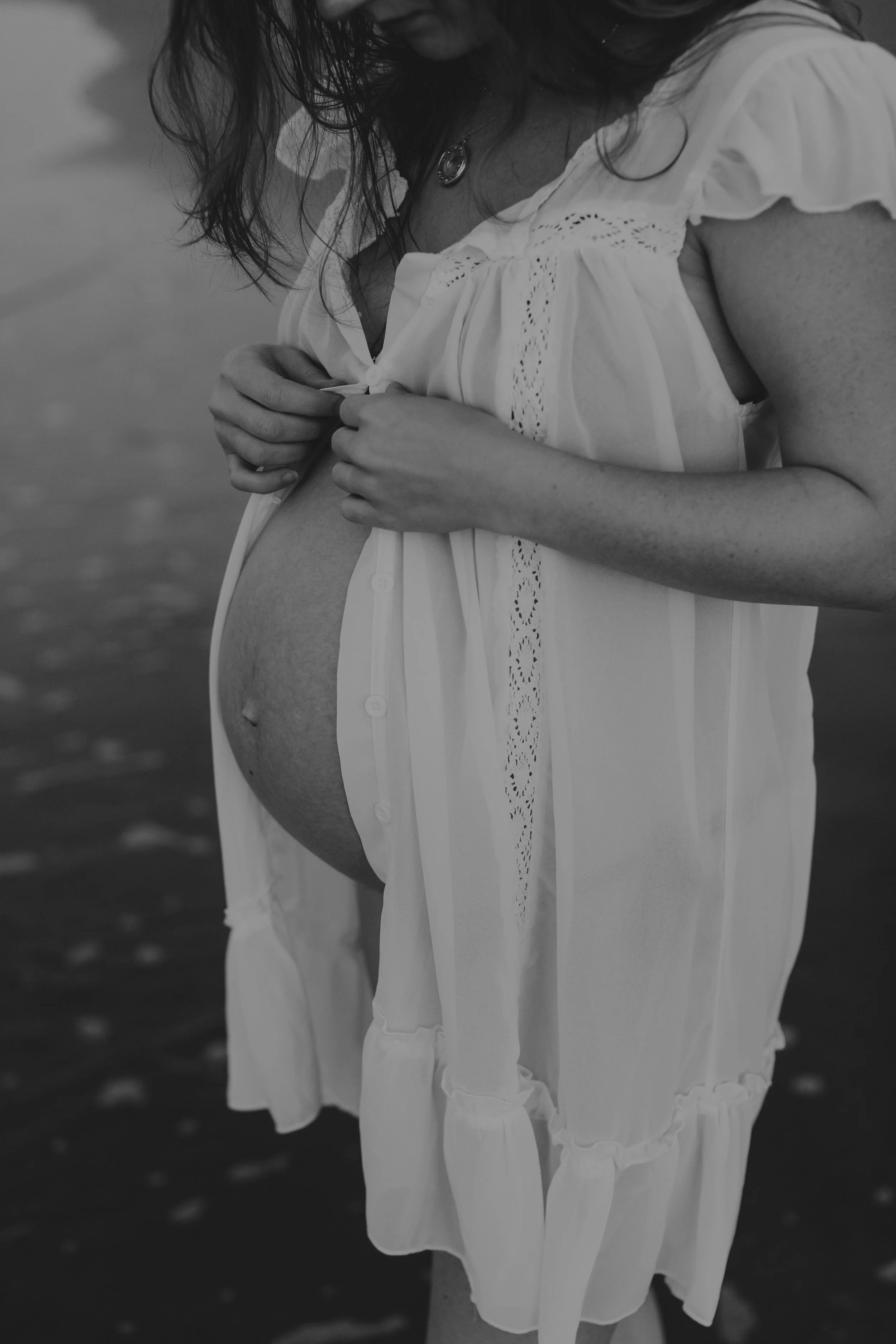 A pregnant woman in a white dress is standing by a body of water, adjusting her dress. The photo is in black and white.