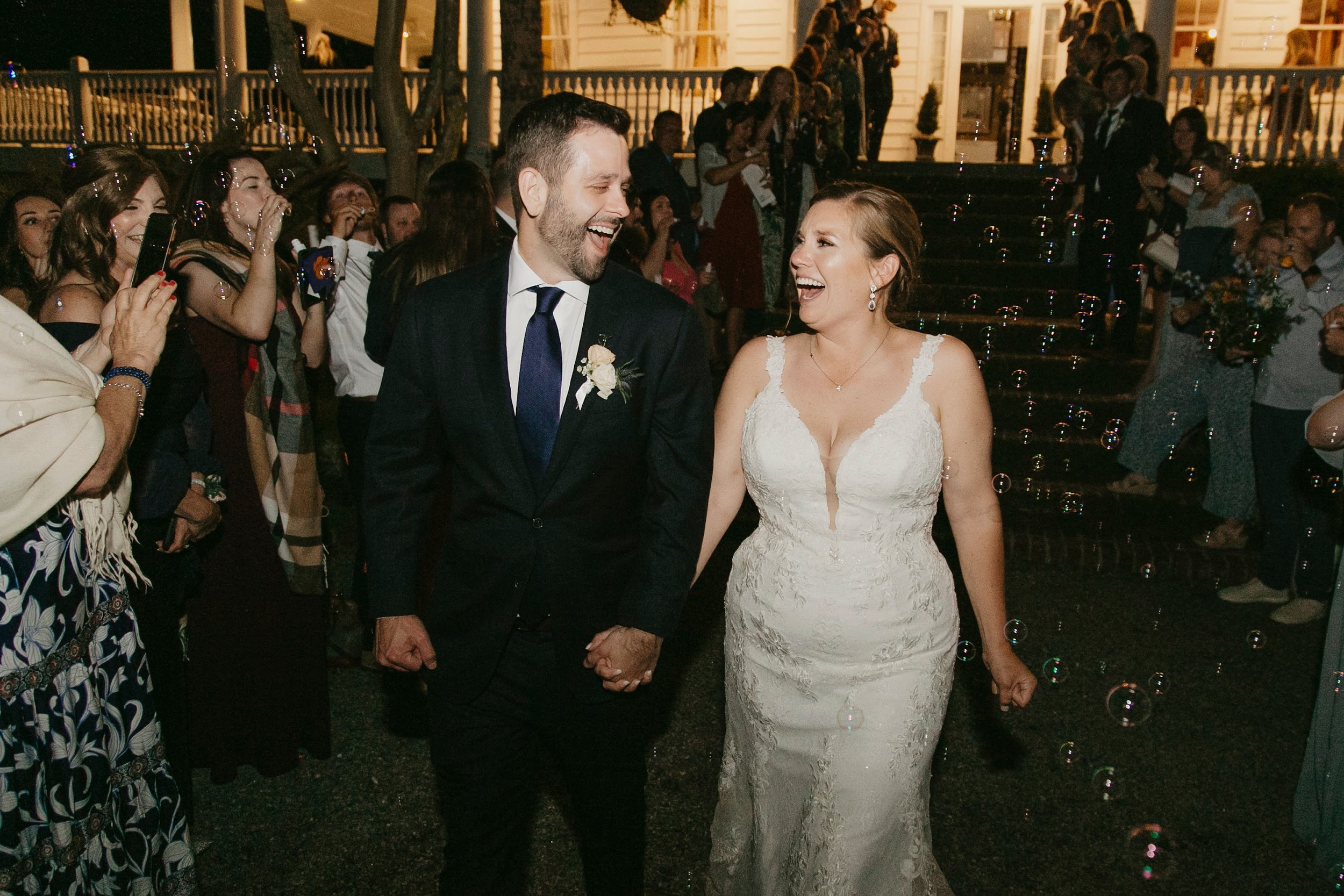 A newly married couple holding hands and smiling at each other during their wedding celebration, surrounded by guests and bubbles in the evening outside house. Old Wide Awake Wedding