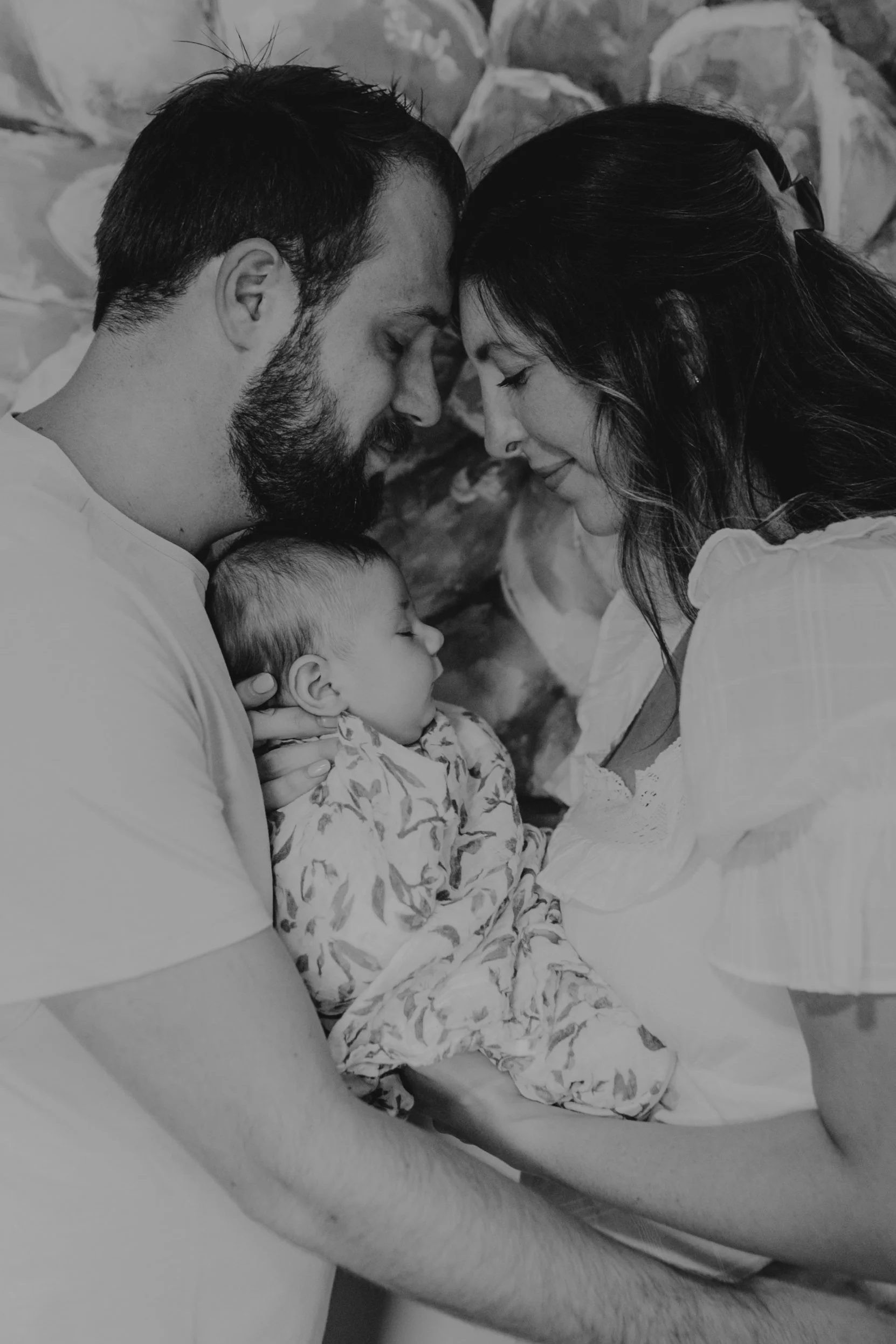 A black and white photo of a family moment with a man, woman, and baby. The man and woman are facing each other with heads touching, the man holding a sleeping baby who is nestled peacefully in his arms. The family appears to be sharing an intimate, loving moment, and the background shows a stone wall.