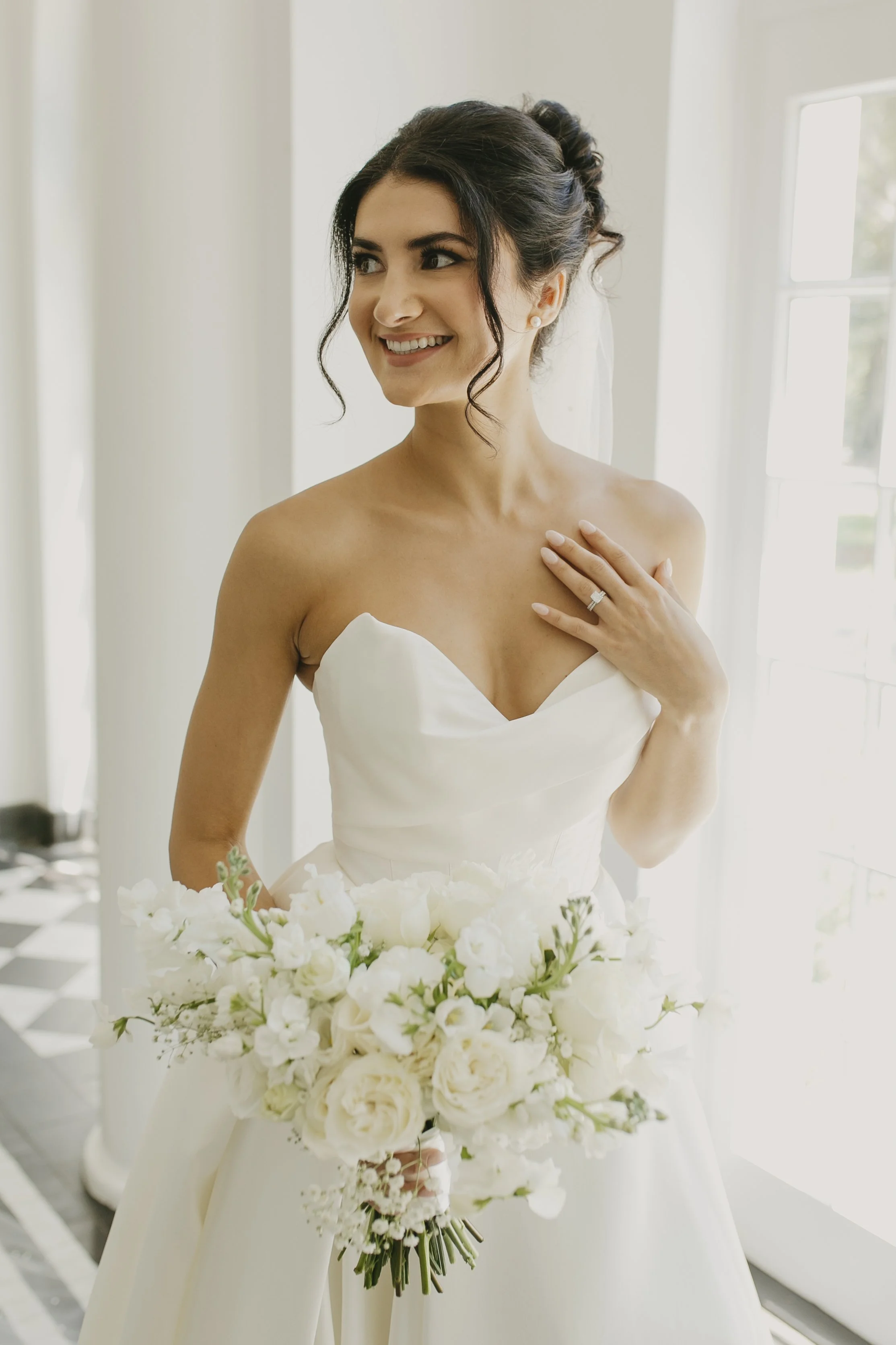 A smiling bride with dark hair in an updo, wearing pearl earrings, holding a bouquet of white flowers in a bright room with large windows