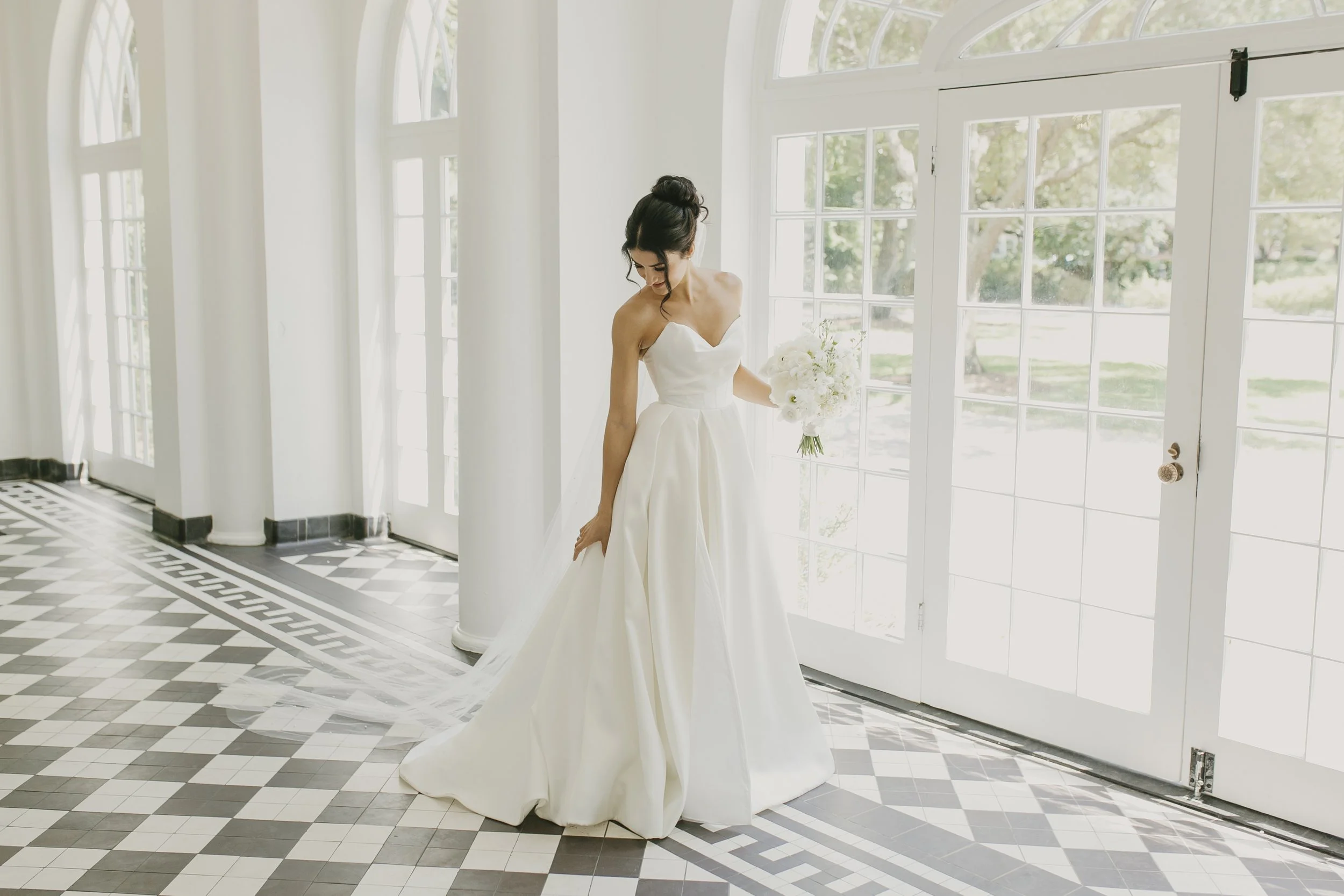A bride in a white wedding dress holding a bouquet, standing inside a bright room with large glass doors and patterned black and white tiled floor.
