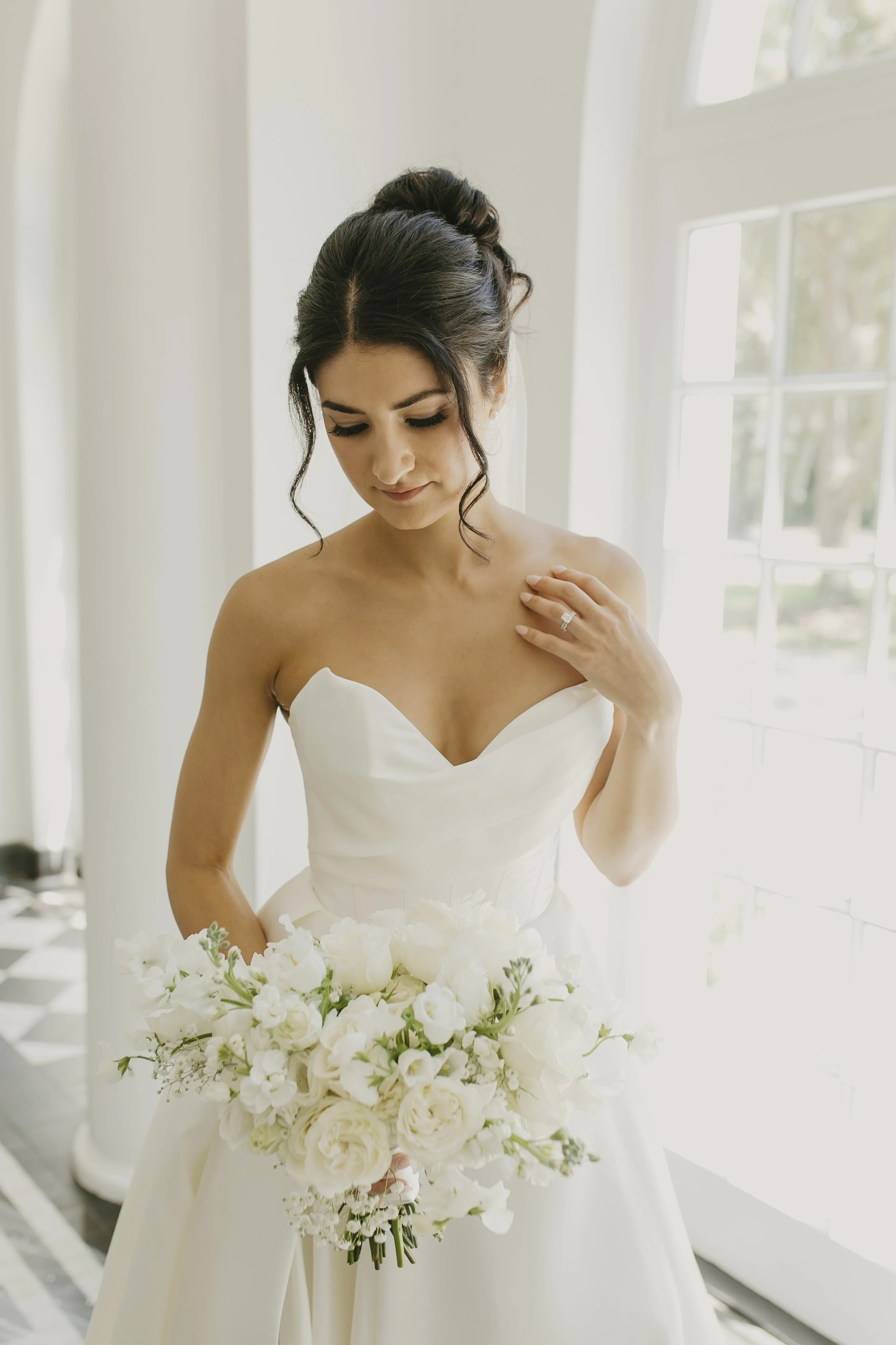 A bride in a strapless white wedding dress holding a bouquet of white flowers inside a bright room with large windows.
