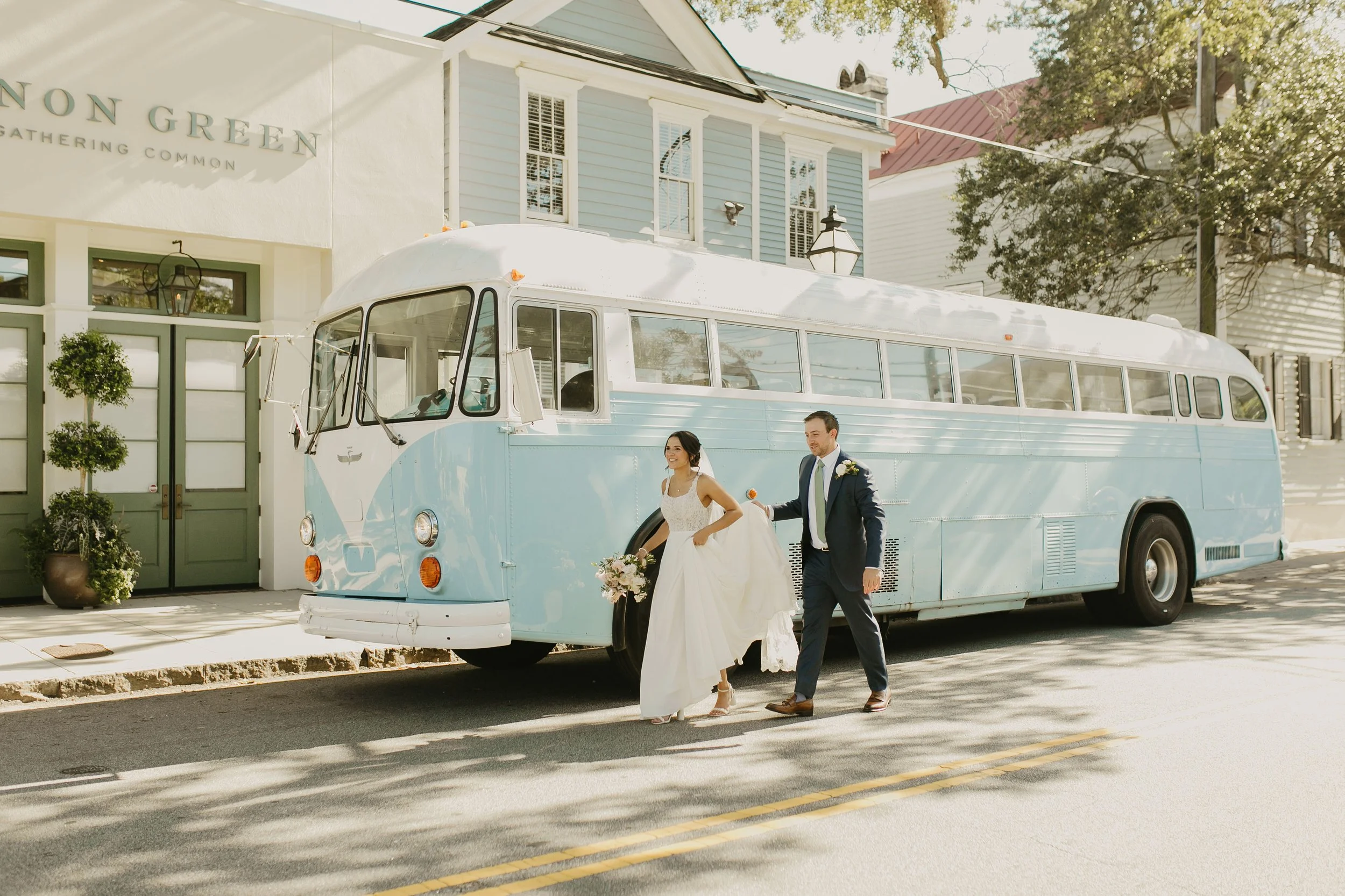 Bride and groom walking away from a vintage blue bus parked on a street, with the bride holding her wedding dress and a bouquet, surrounded by trees and buildings in a sunny daytime setting. Cannon Green Wedding