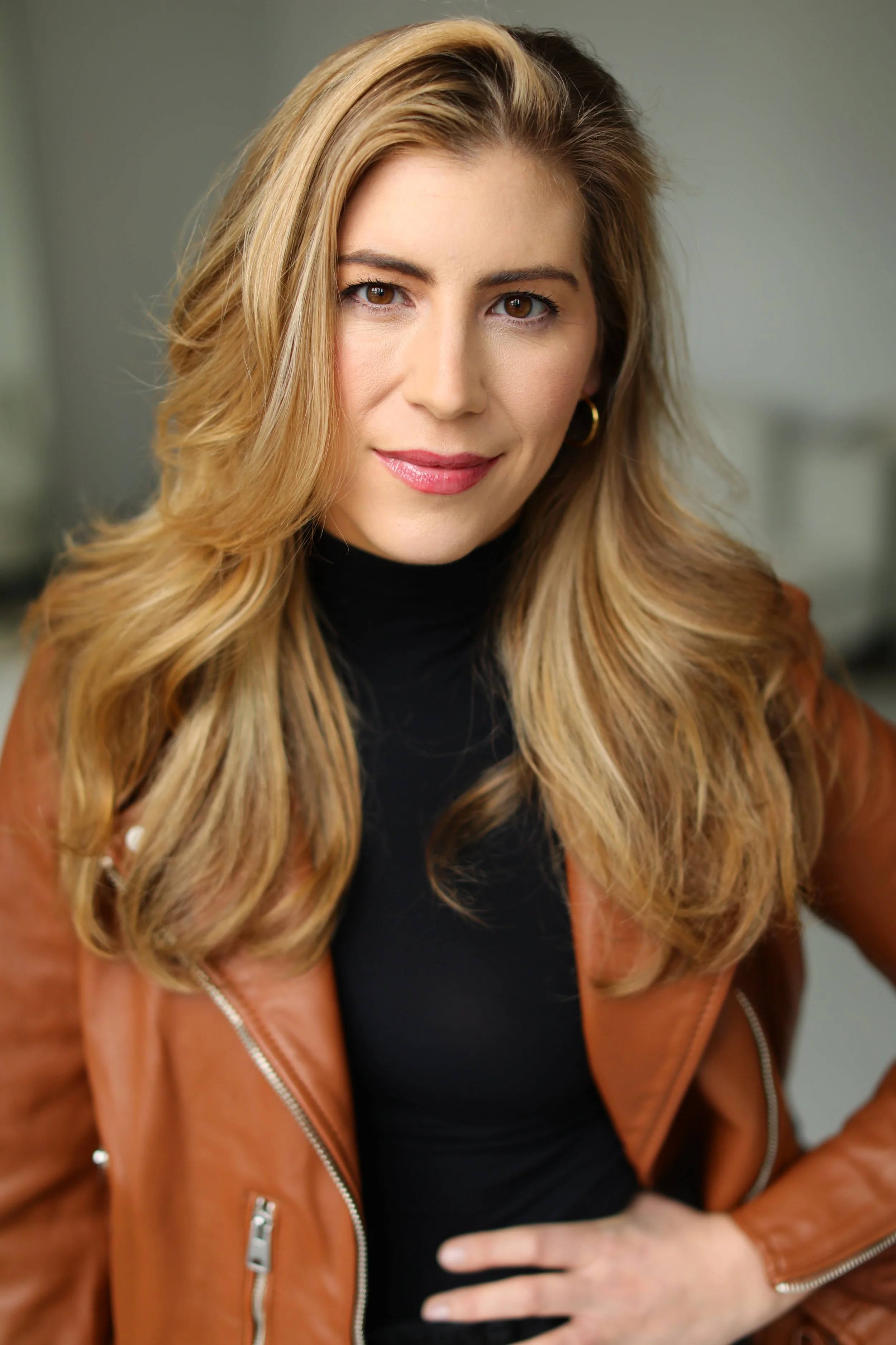 A woman with long wavy blonde hair wearing a black turtleneck, brown leather jacket, and gold hoop earrings, smiling in front of a neutral background.
