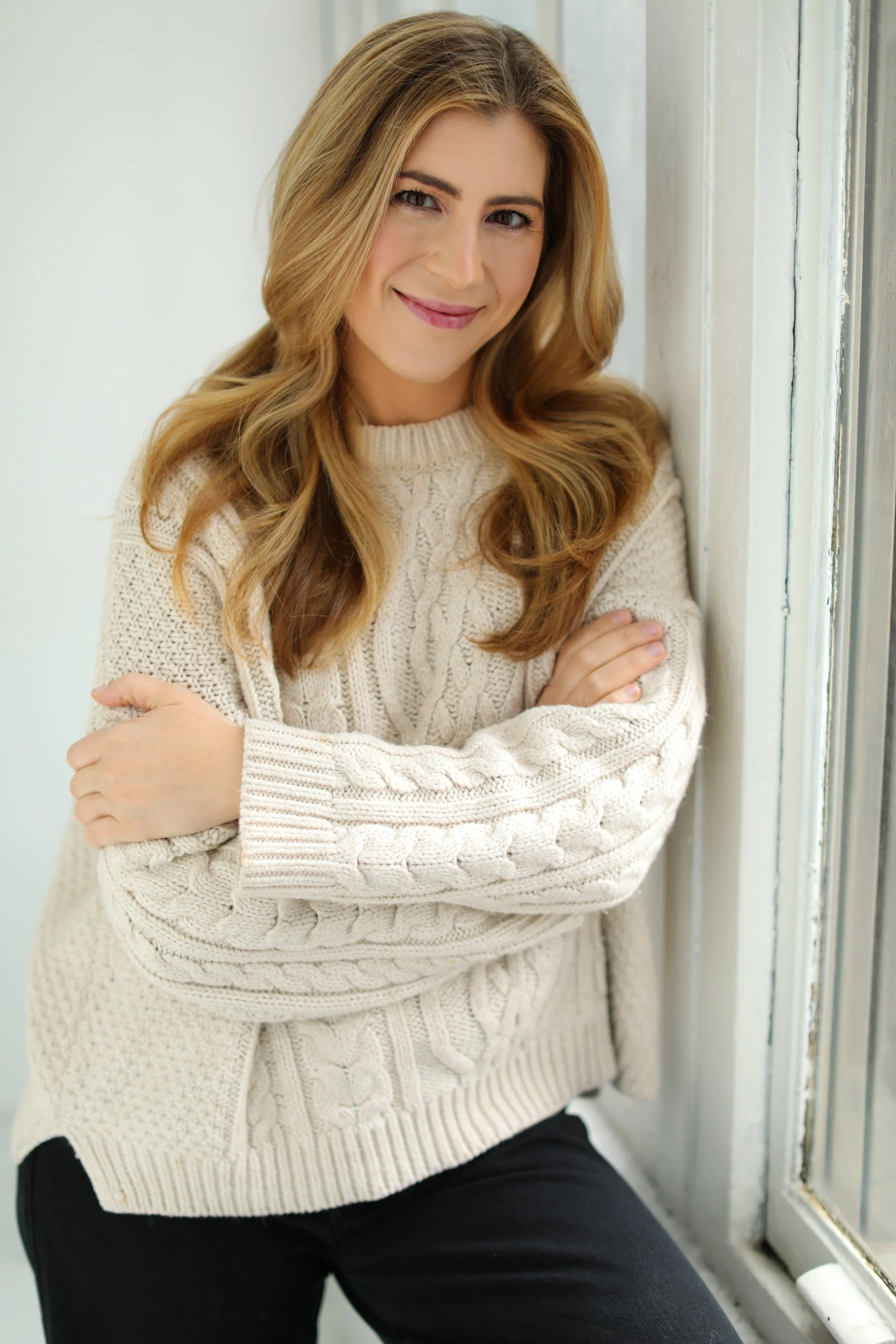 A woman with long blonde hair smiling, wearing a cream cable-knit sweater and black pants, sitting by a window.