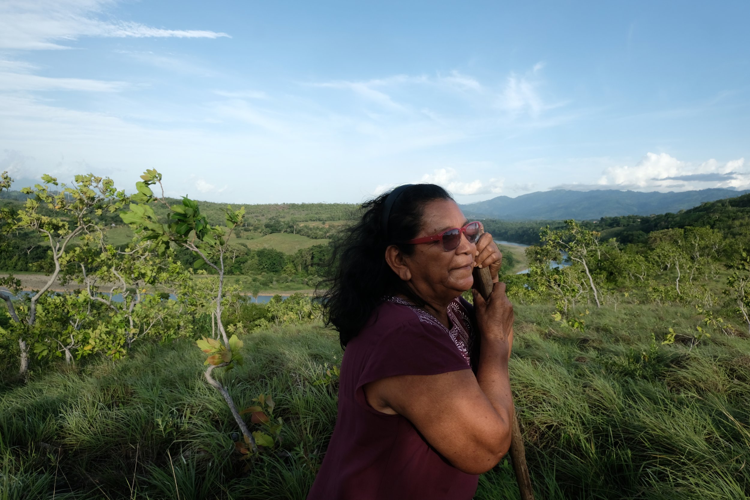 Mujer de cabello oscuro y gafas rojas, con expresión pensativa, en un paisaje rural con árboles verdes, río y montañas al fondo bajo cielo azul con nubes.