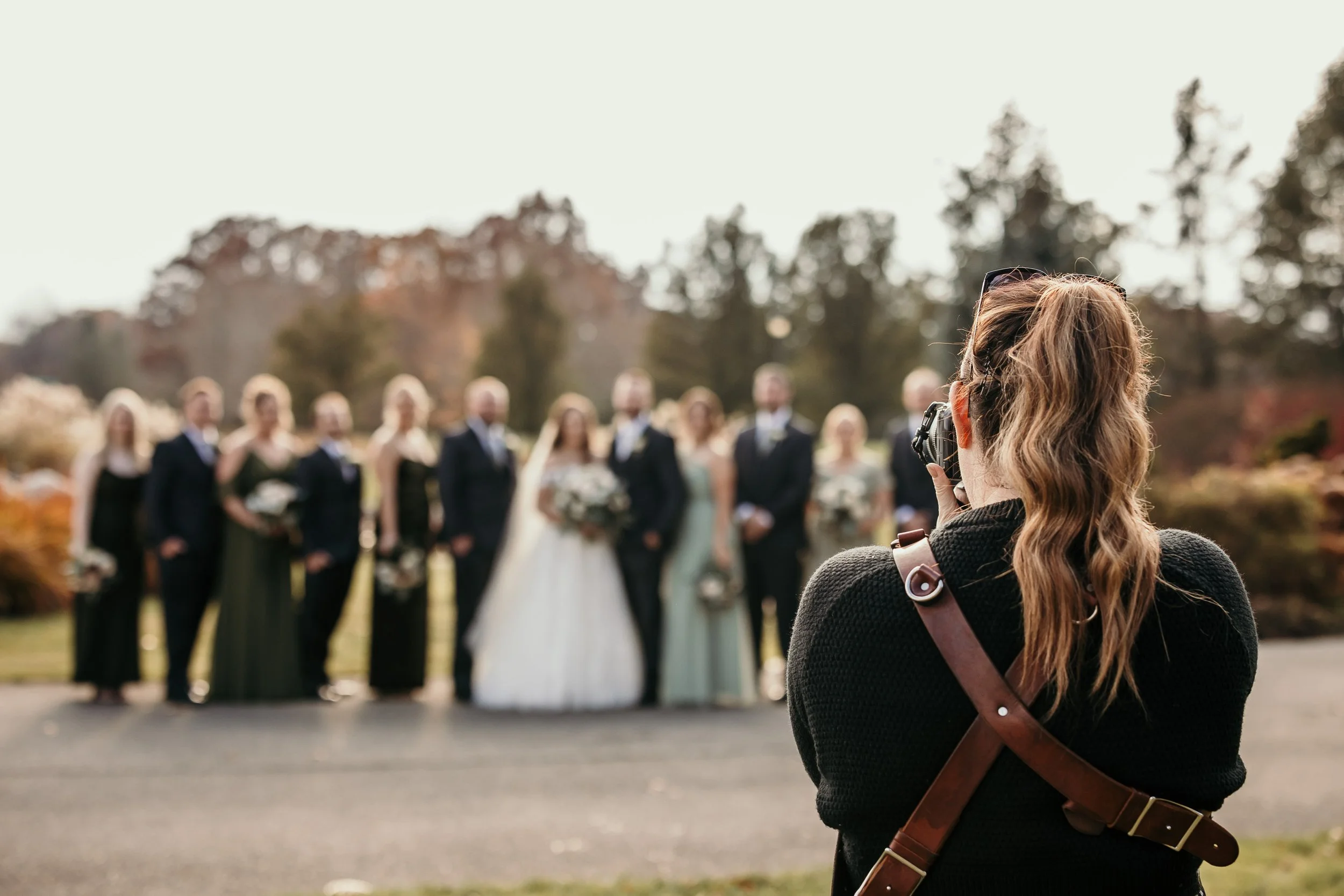 A woman with long hair and a dark sweater taking a photo of a wedding party outdoors with a camera. The wedding party includes multiple people dressed in formal attire, standing in a line, with a bride in a white dress holding a bouquet and several groomsmen in suits. The background features trees and an overcast sky.
