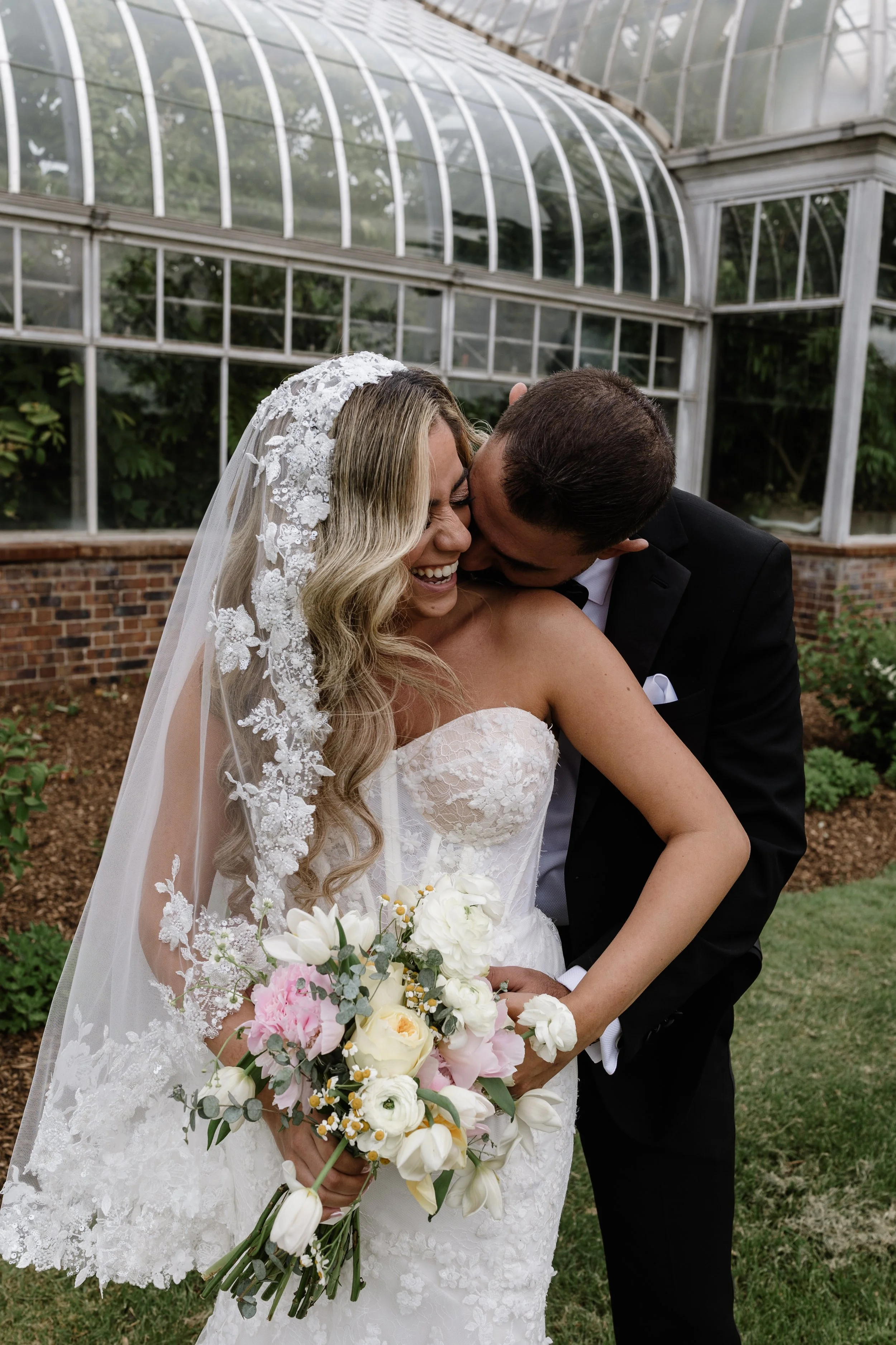 A bride in a white lace wedding dress and veil holds a bouquet of white, pink, and yellow flowers. A groom in a black tuxedo is kissing her cheek inside a greenhouse with glass panes and greenery visible behind.