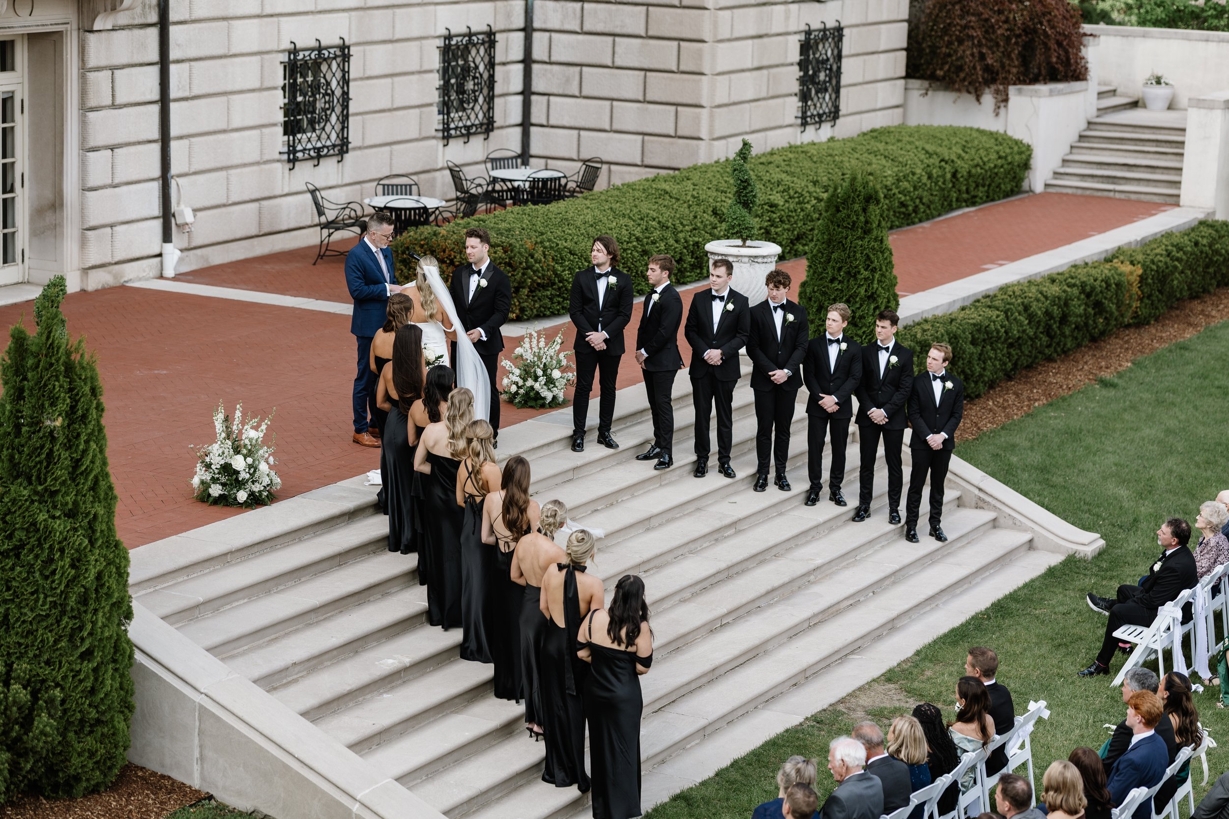 A wedding ceremony taking place outdoors on steps in front of a large stone building. The bride and groom stand facing each other, with the officiant reading from a paper. The bride is dressed in white and wearing a veil. The groom and groomsmen are in black tuxedos with white shirts and black bowties. Bridesmaids and guests are seated, watching the ceremony. The area is decorated with floral arrangements and greenery.