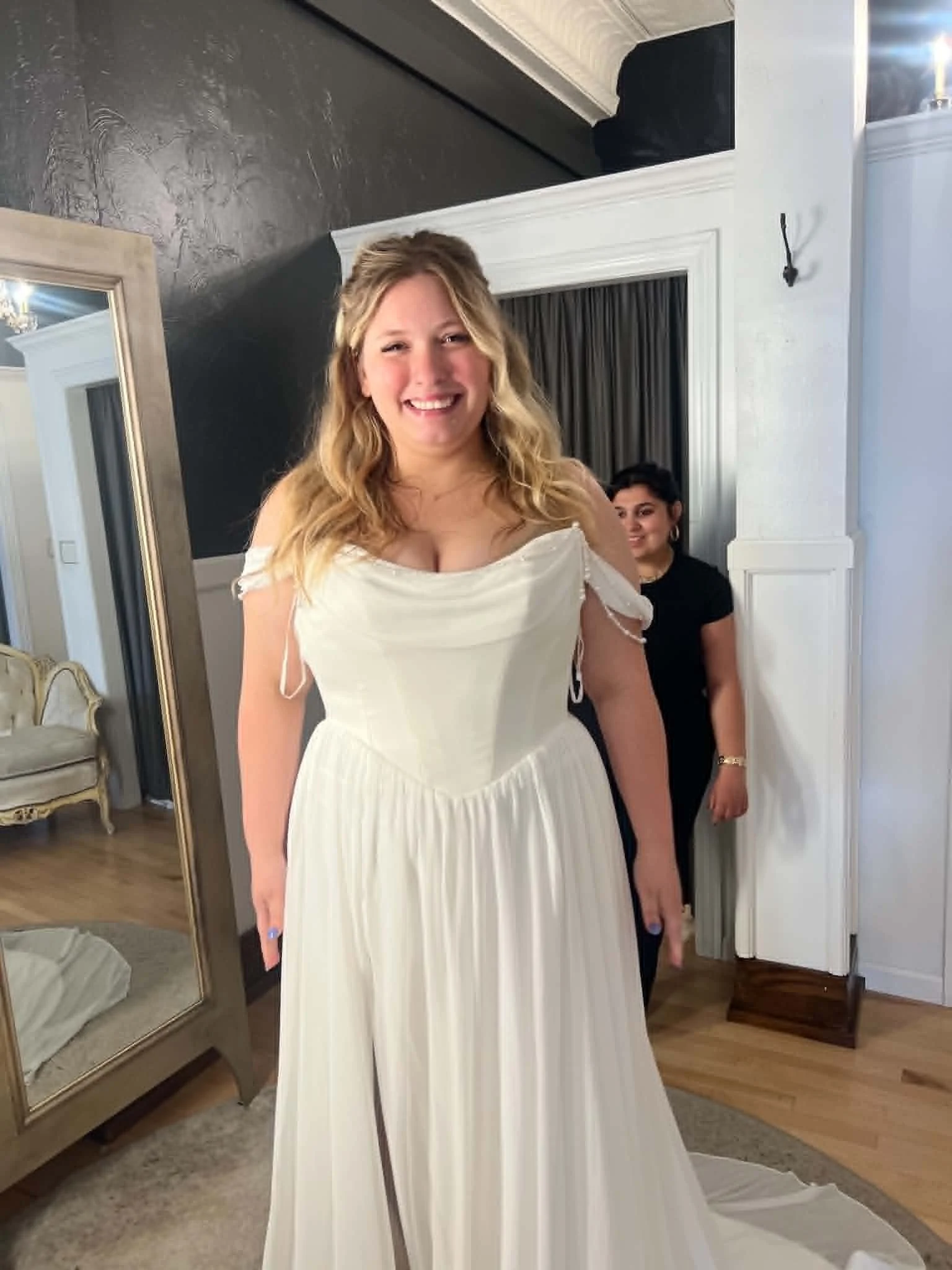 A woman in a white dress is smiling in front of a mirror in a room. There is a young girl in the background wearing dark clothing, standing near a white wall with a dark curtain. The room has wooden floors and a mix of white and dark-colored walls.