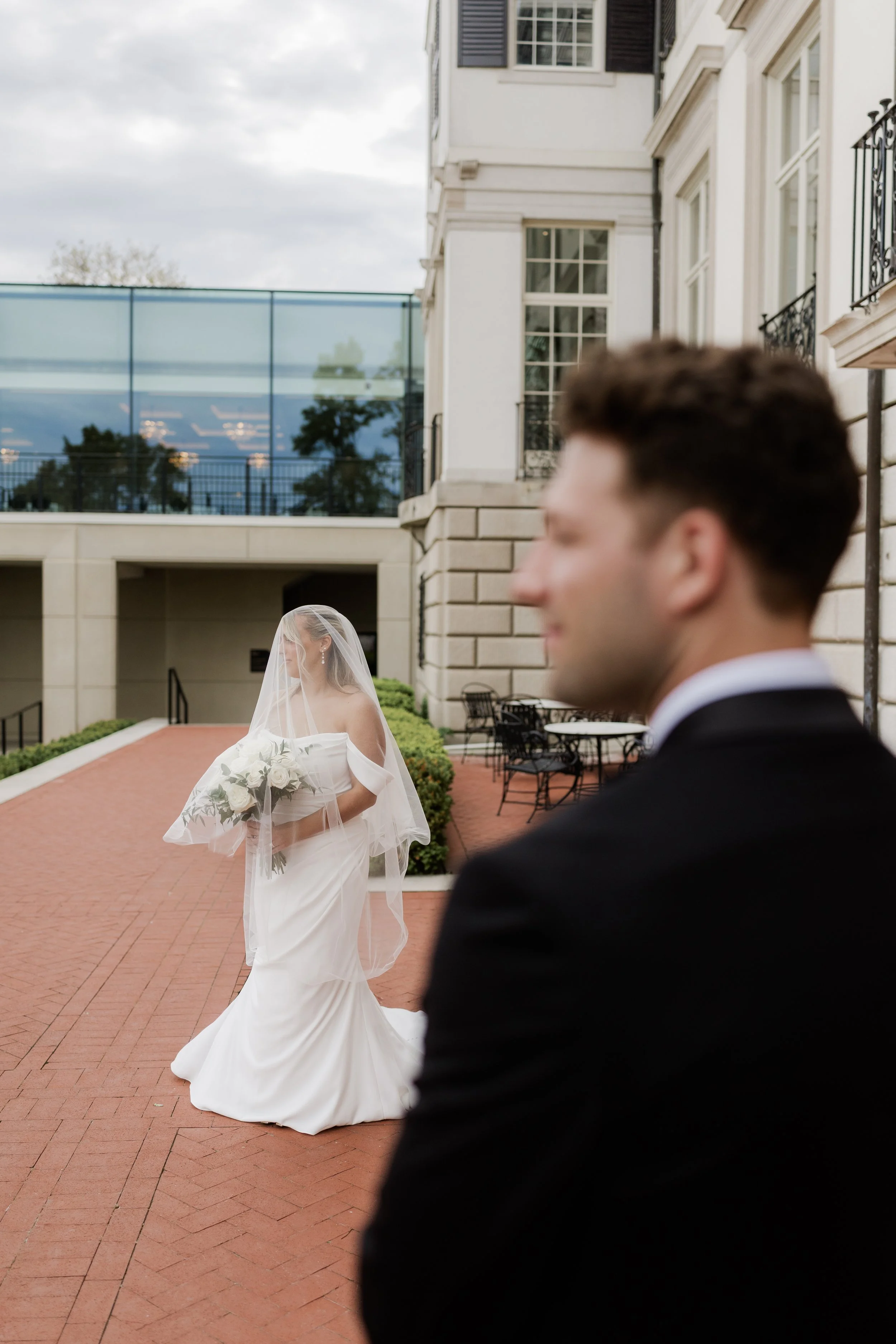 A bride in a white wedding dress carrying a bouquet of white flowers walks outside, with a groom in a black tuxedo in the foreground, blurred, during a wedding ceremony.
