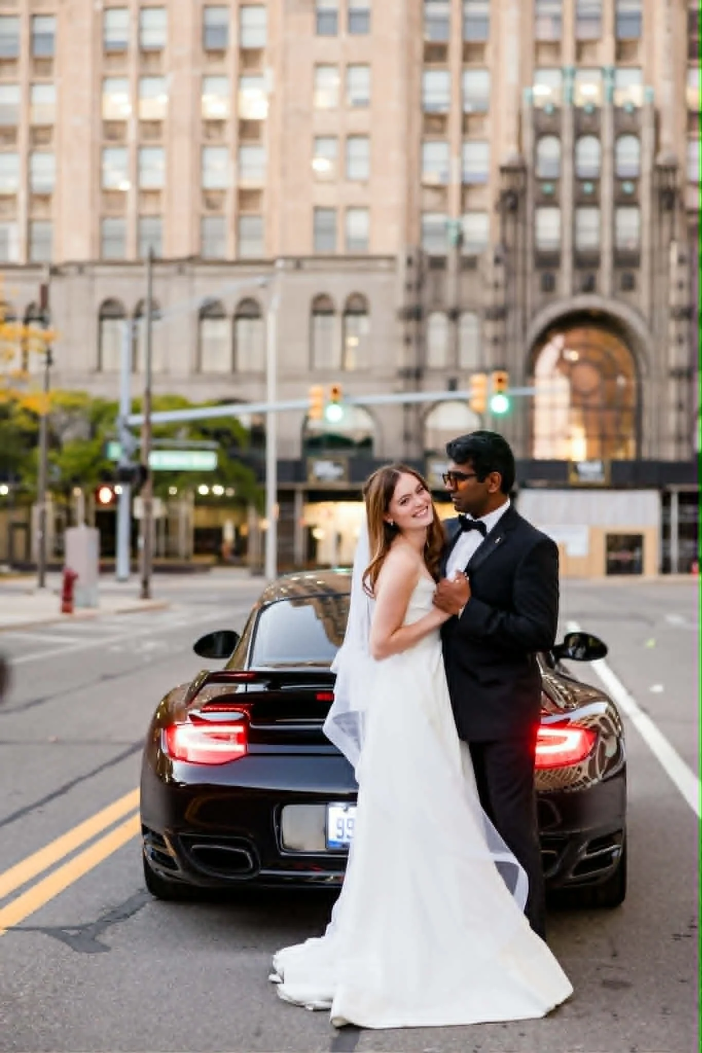 A bride and groom standing embrace in front of a black luxury car, with a historical building in the background