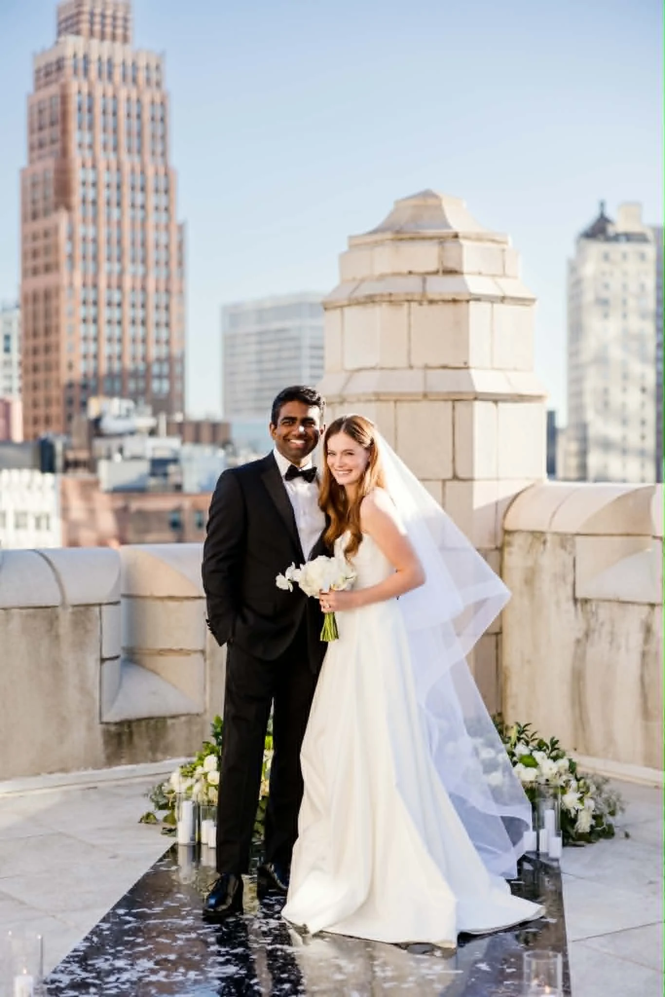 A newlywed couple on their wedding day standing on a rooftop terrace in an urban setting with tall buildings in the background. The bride is holding a bouquet of white flowers, wearing a white wedding gown and veil, and the groom is dressed in a black tuxedo. They are smiling and standing close together, surrounded by floral arrangements and candles on the floor.