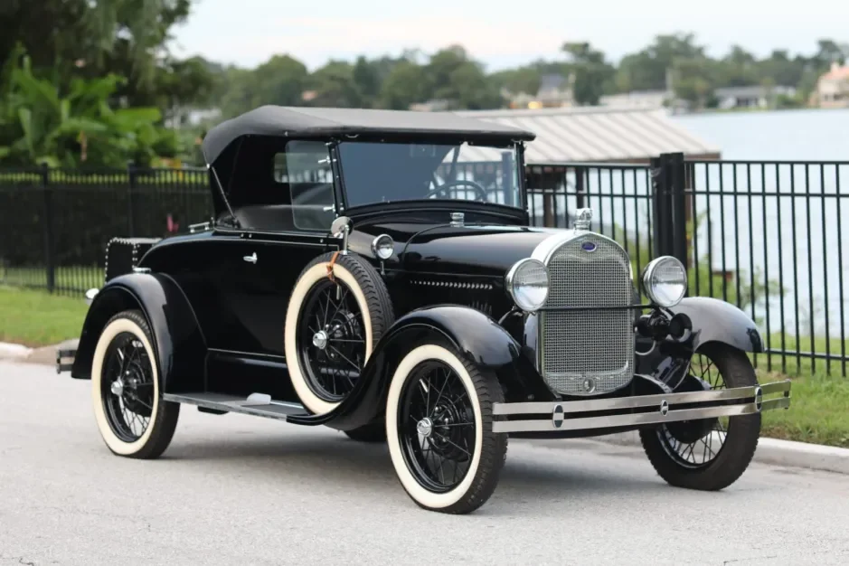 A vintage black car with whitewall tires parked on a paved road near a fence, with trees and water in the background.