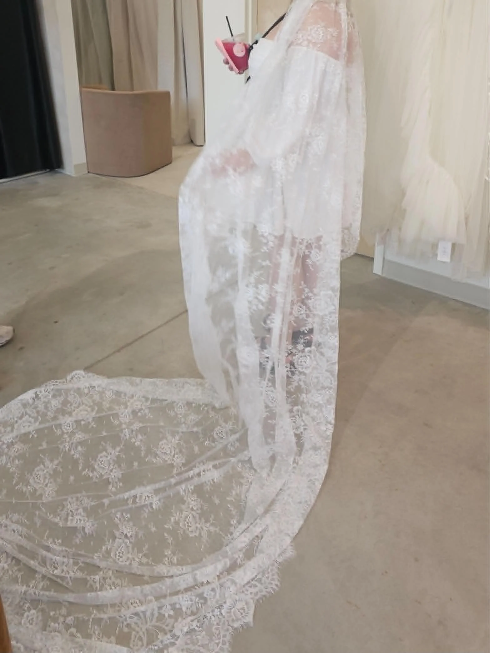 A person wearing a dress is taking a mirror selfie with a smartphone. The dress is white with lace details, and they are holding a cup with a straw. The setting appears to be a room with a carpeted floor, a beige chair, and curtains in the background.