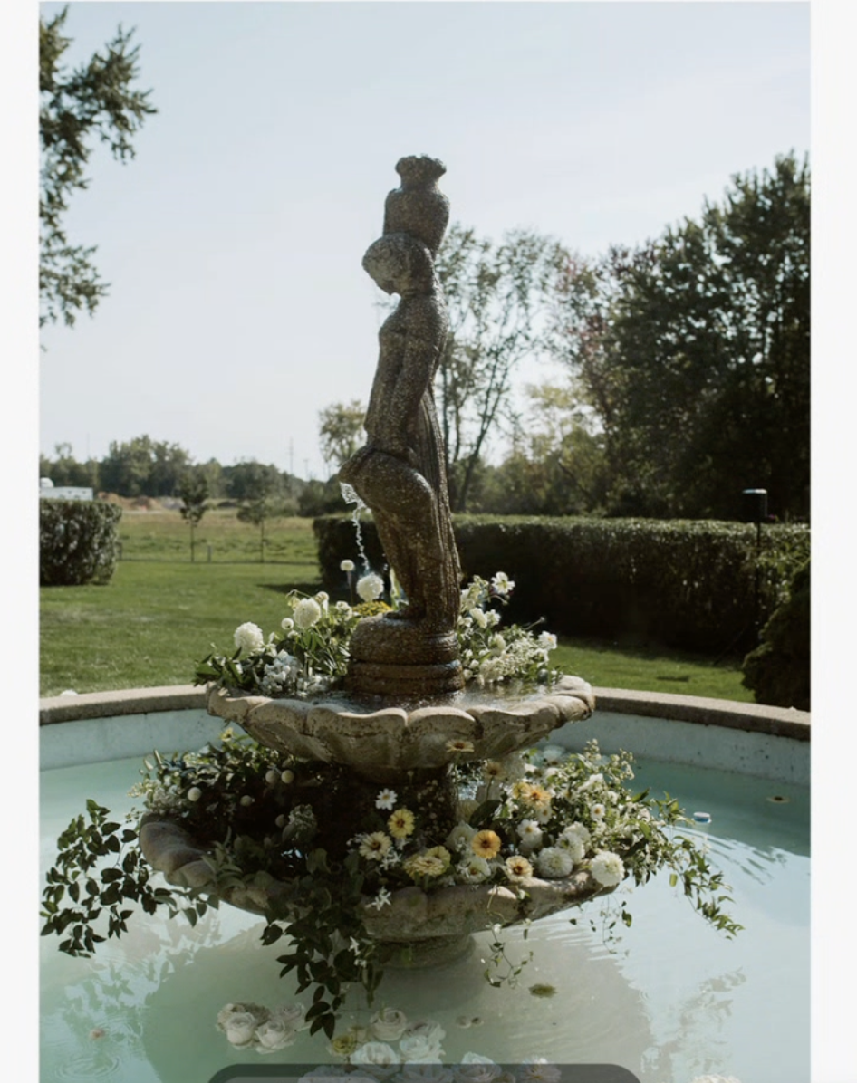 A stone fountain featuring a sculpture of a girl holding a vase, surrounded by flowers, in a garden with trees and lawns in the background.