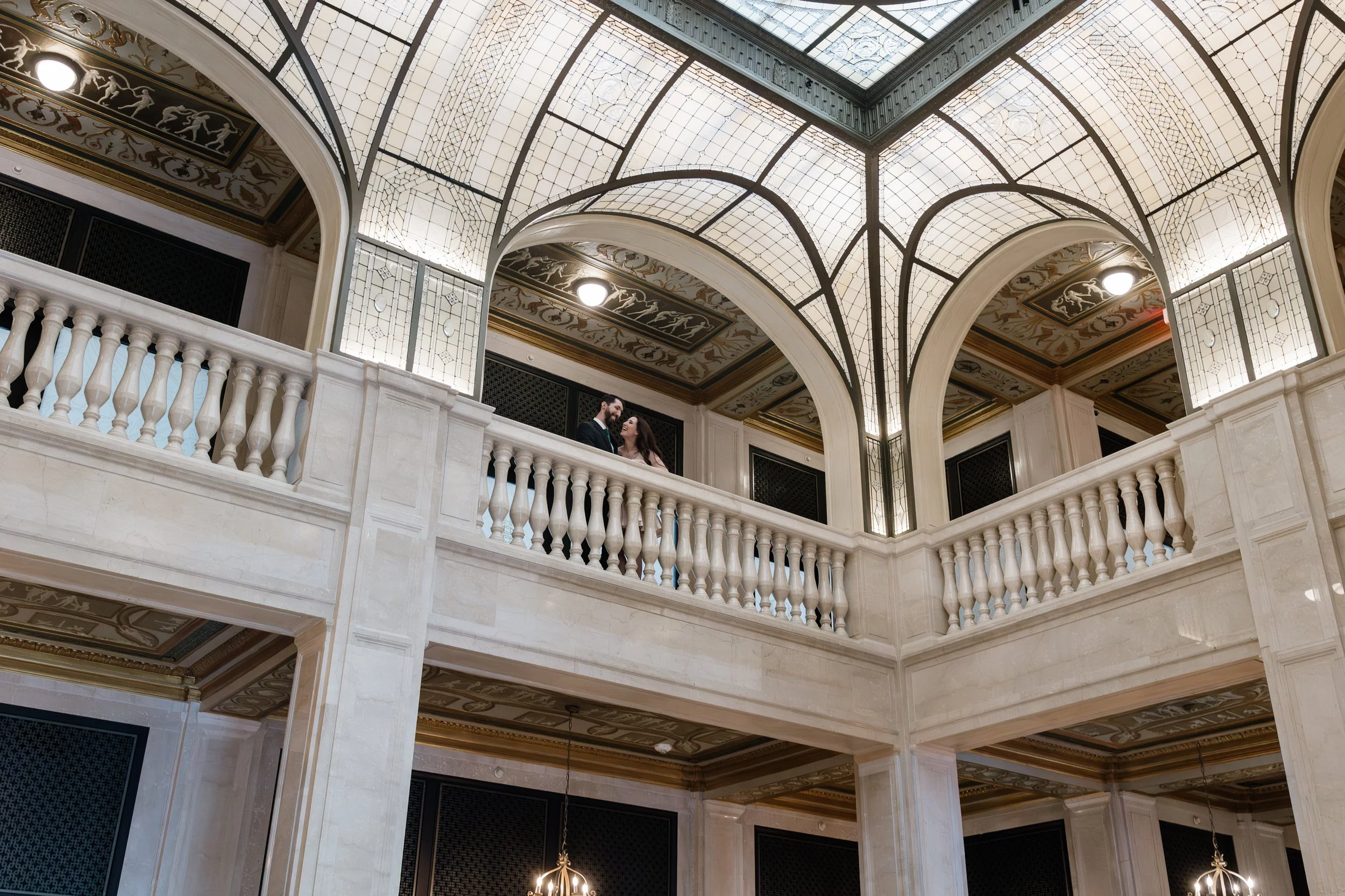 Photo of an elegant, historic interior lobby with a high, ornate ceiling featuring stained glass and gold accents. A balcony with a white balustrade overlooks the space, where a couple in formal attire stands, smiling at each other.