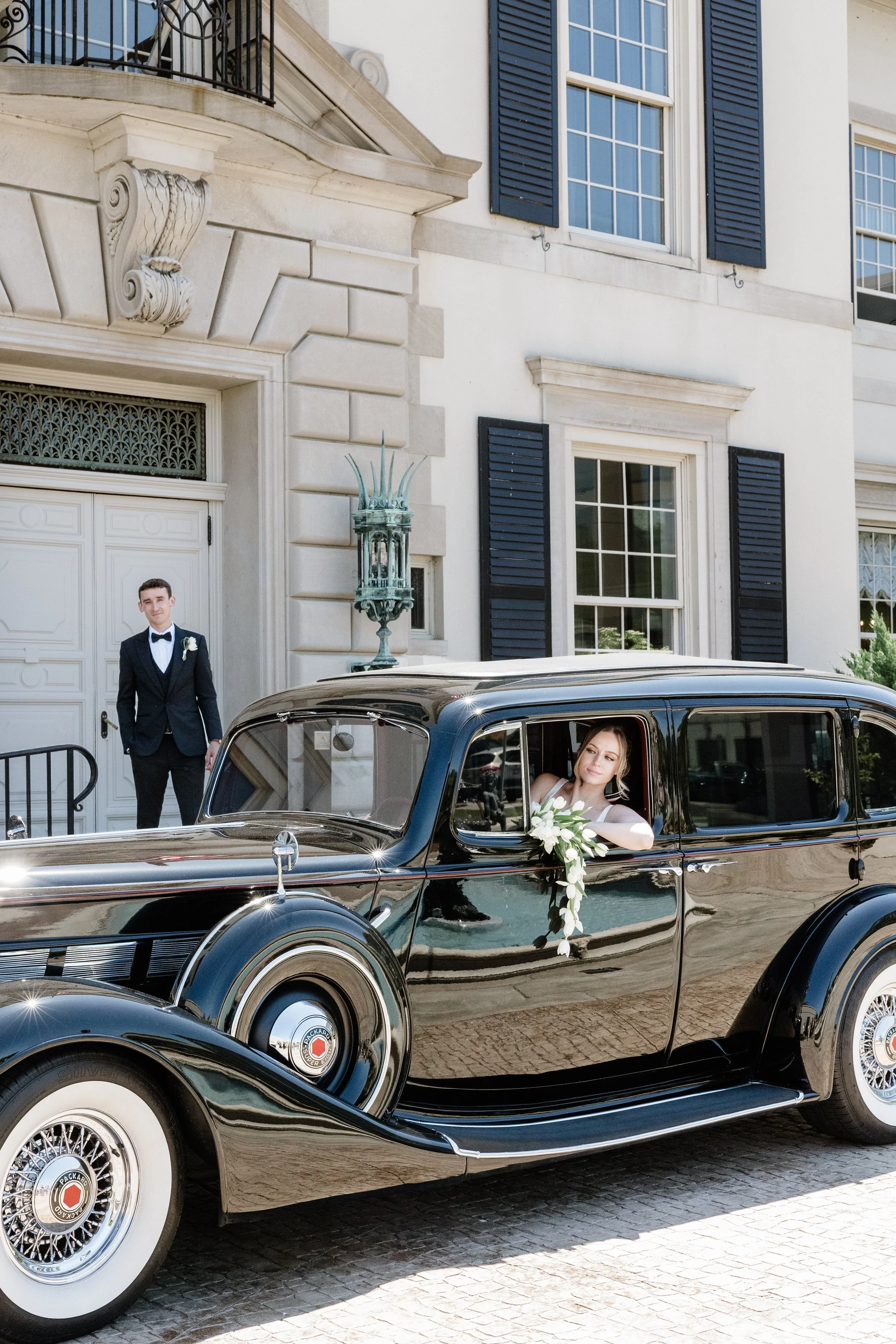 A woman sitting inside a black vintage car holding white flowers, with a man in a tuxedo standing outside in front of a mansion with large windows and black shutters.