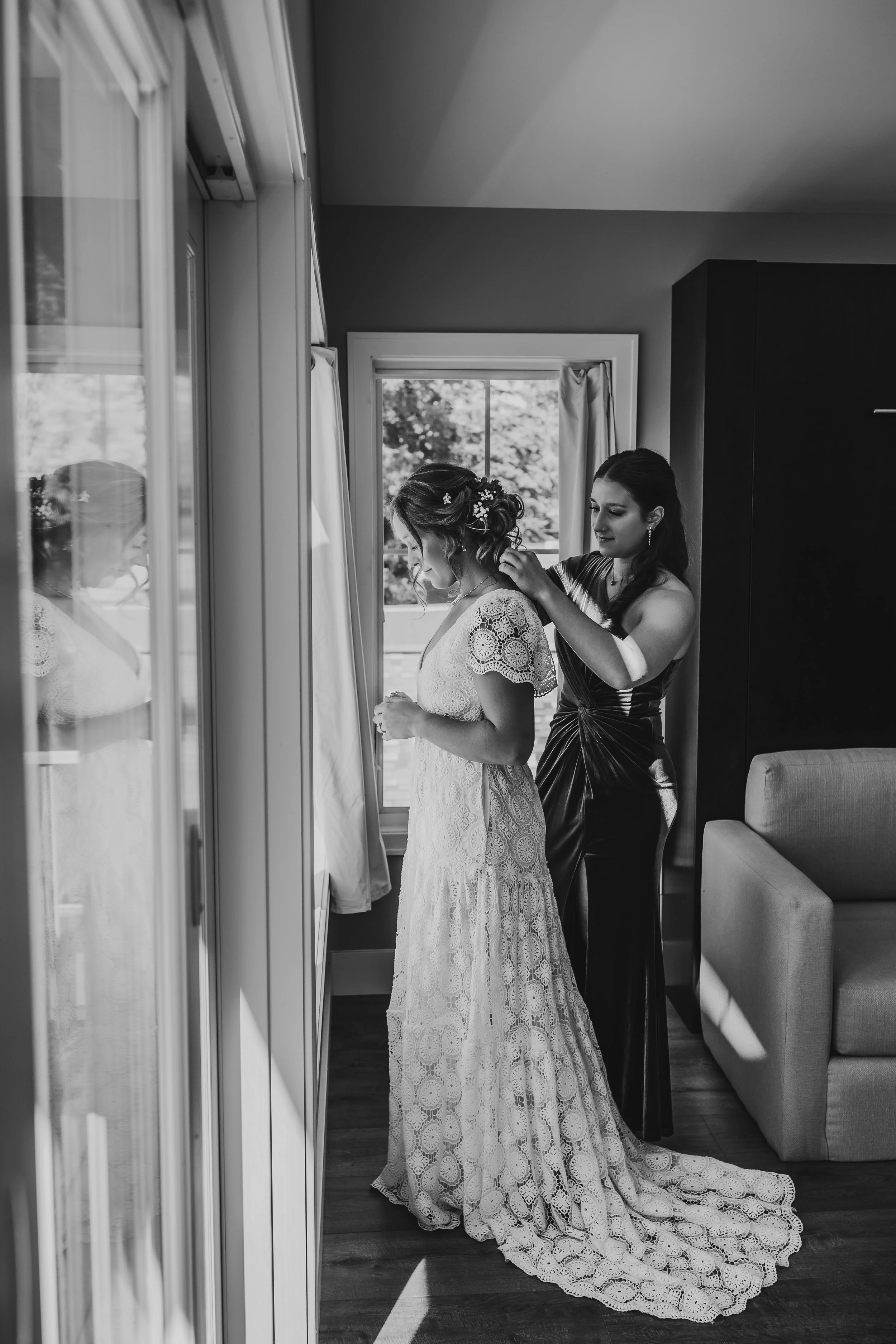 Black-and-white photo of a bridesmaid helping the bride with her necklace, captured in soft window light with a reflection adding depth to the intimate moment.