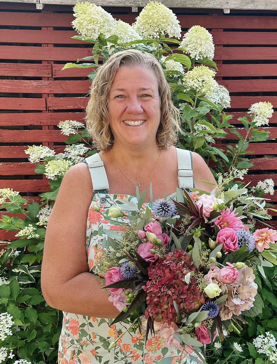 A woman with curly blonde hair smiling while holding a large bouquet of colorful flowers in front of a backdrop of white hydrangeas and a red wooden fence.