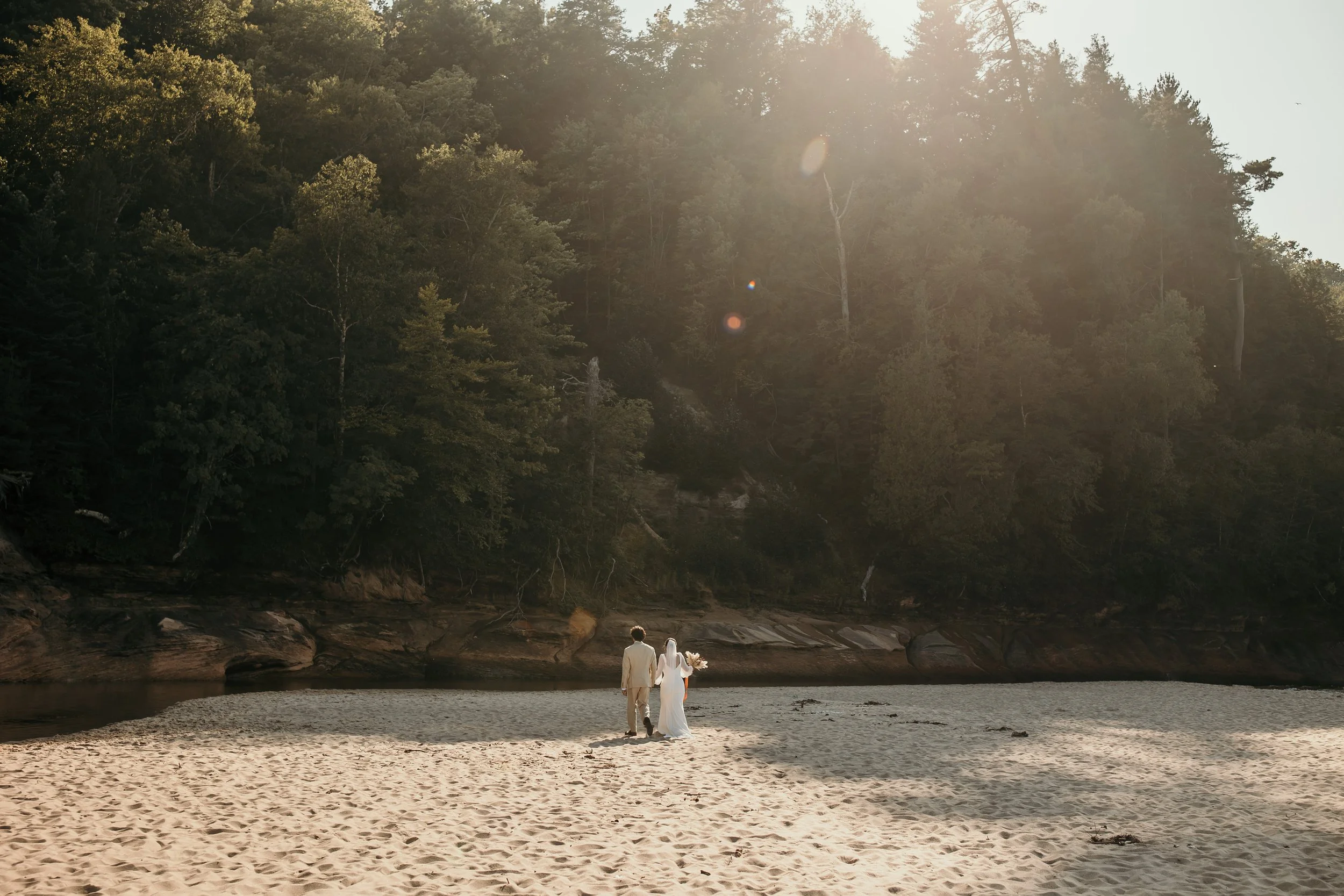 A bride and groom walk hand-in-hand on a sandy beach at sunset, with a dense forest and tall trees in the background.