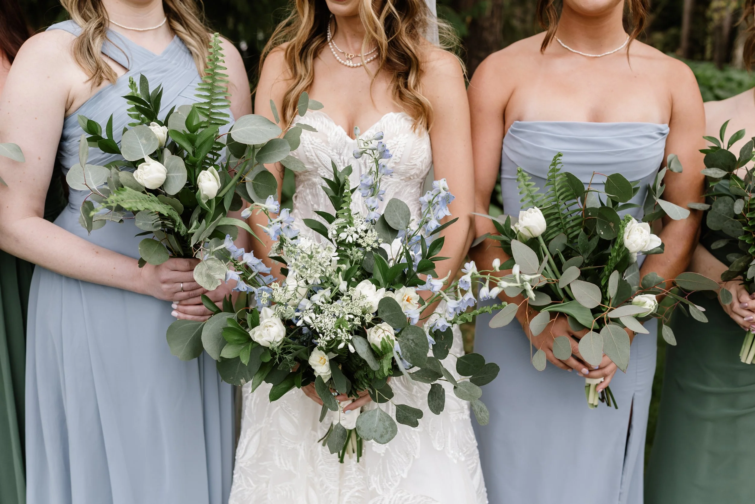 Three women dressed in beige and light-colored dresses, with the middle woman wearing a white lace dress, standing behind a large floral arrangement with white and cream-colored roses and green foliage.