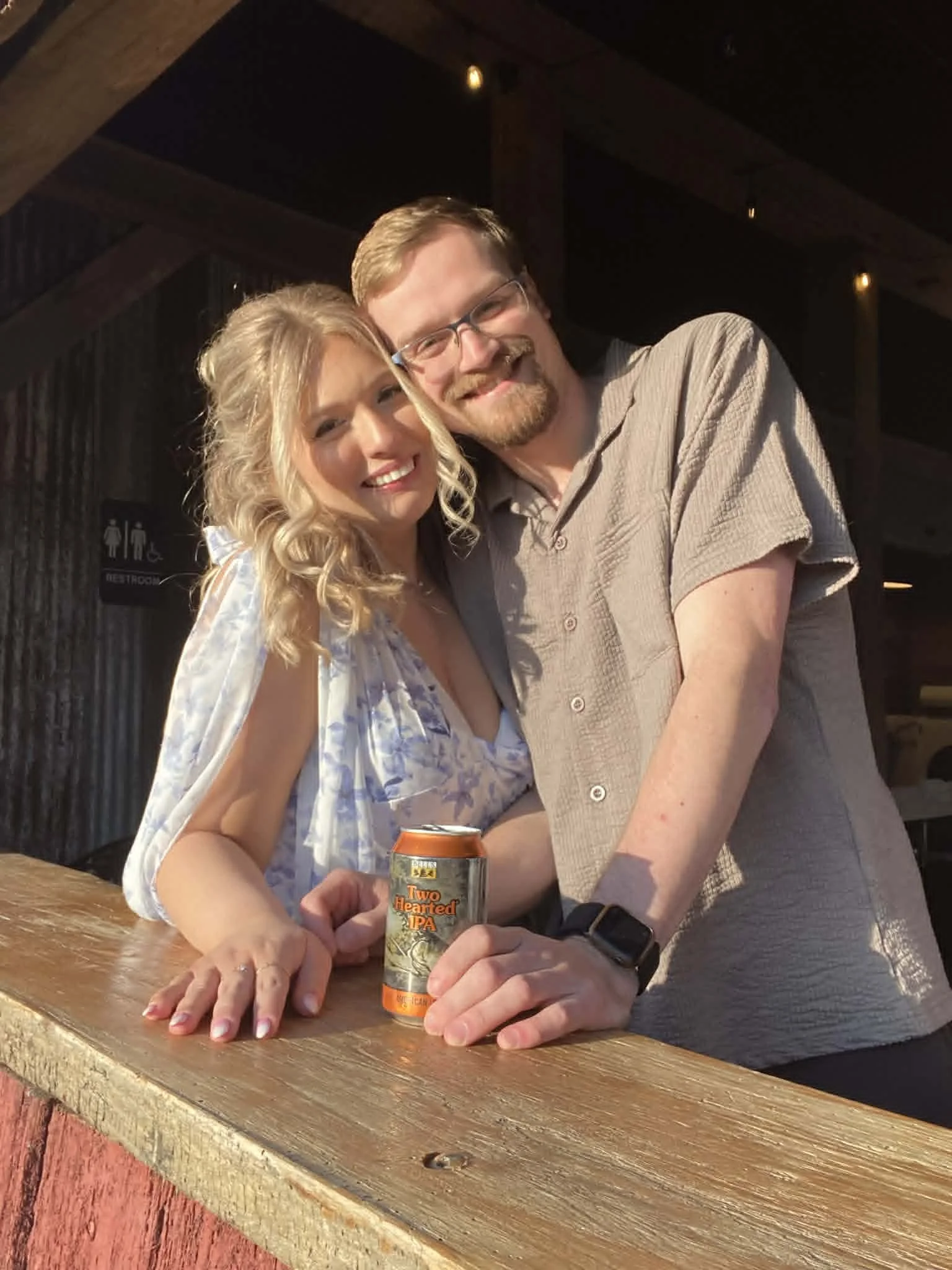 A smiling couple at a bar with the woman holding a can of beer labeled 'Two Hearted IPA' and the man's arm around her. They are inside a dimly lit venue with a wooden bar counter.