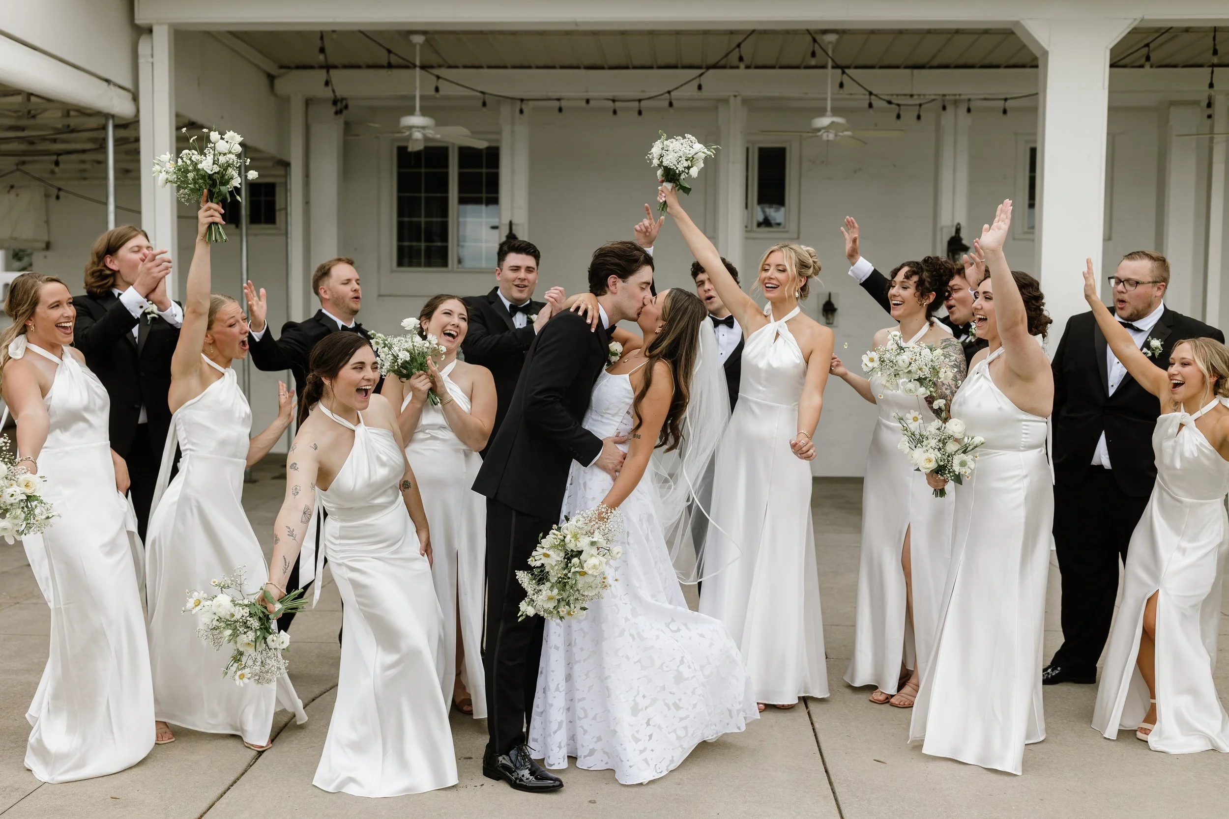 A group of people dressed in wedding attire celebrating outdoors, with a newlywed couple kissing in the center surrounded by bridesmaids and groomsmen holding flowers and raising their hands in celebration.