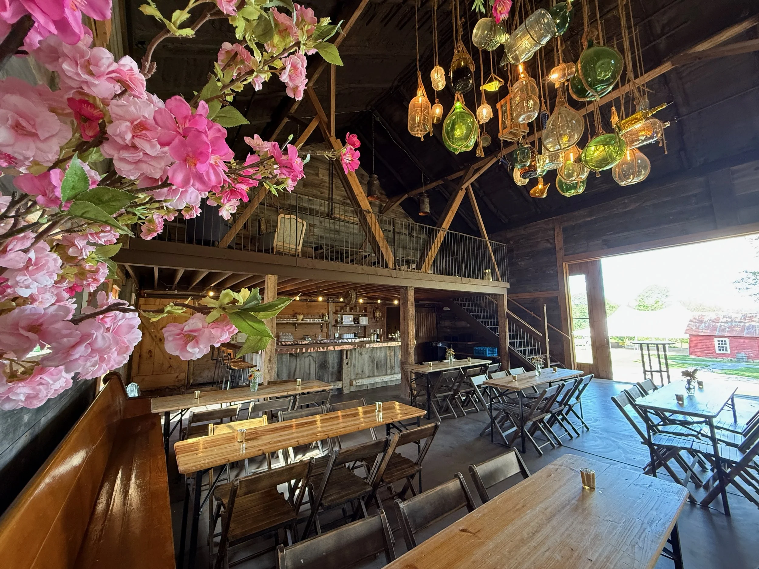Interior of the rustic Brewery Barn with wooden tables and chairs, decorative hanging glass jugs on the ceiling, pink flowers in the foreground, and large open doors letting in natural light.