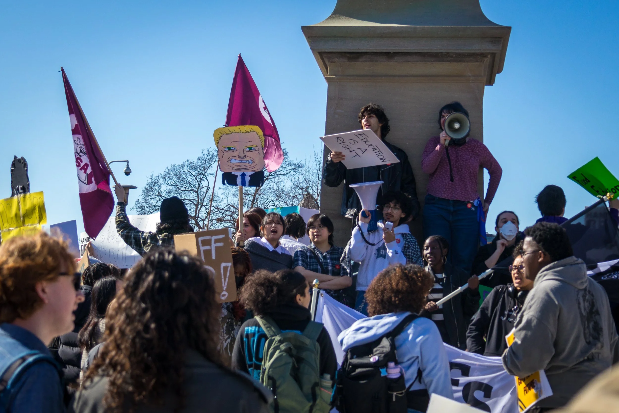 Students speak to a crowd during a rally about OSAP student funding, standing on the base of a monument with a megaphone as protesters hold signs and flags, including one reading “Education Is a Right.”