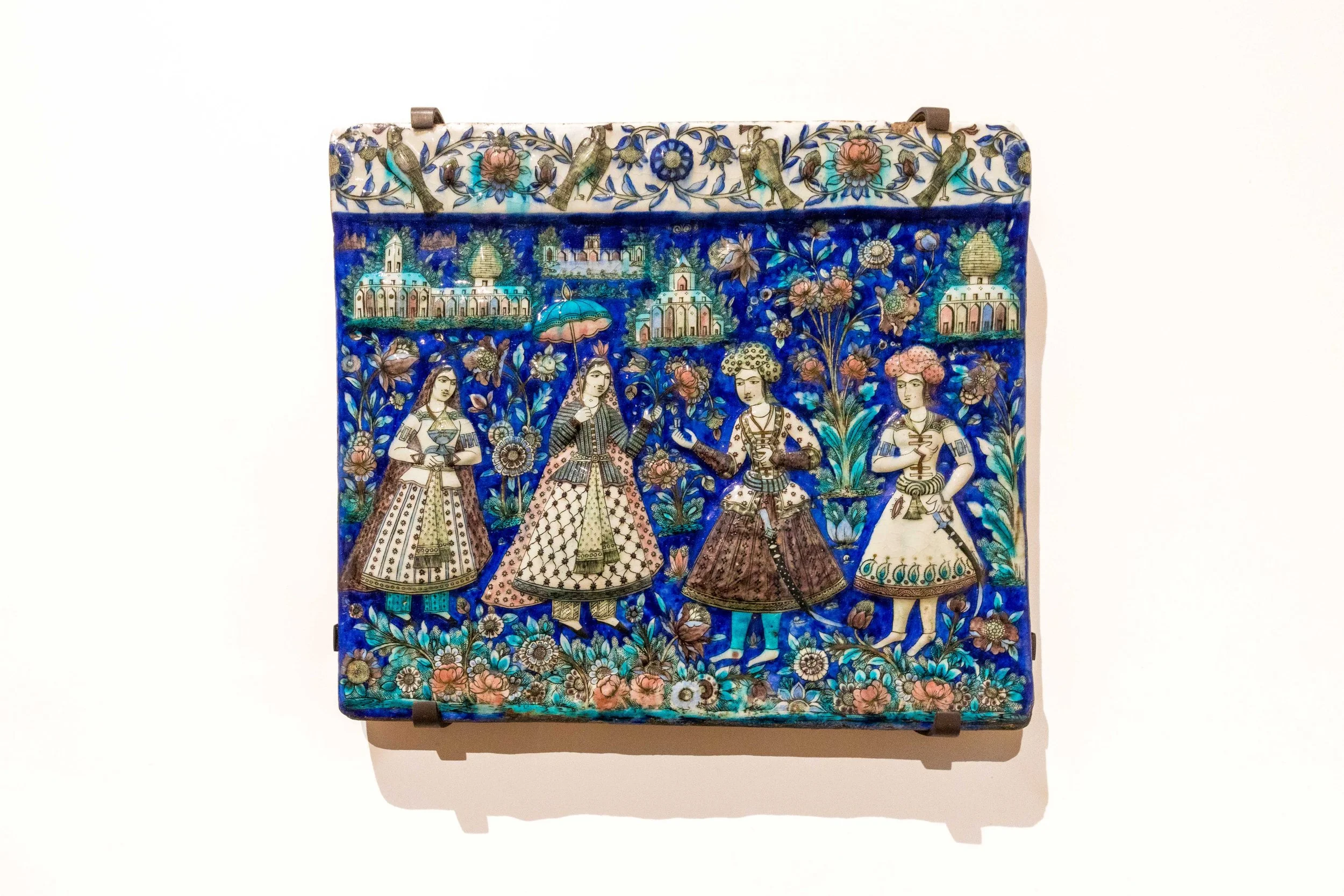 19th-century Qajar-era ceramic tile panel from Iran featuring four elegantly dressed couples surrounded by floral and architectural motifs in vivid cobalt blue tones.