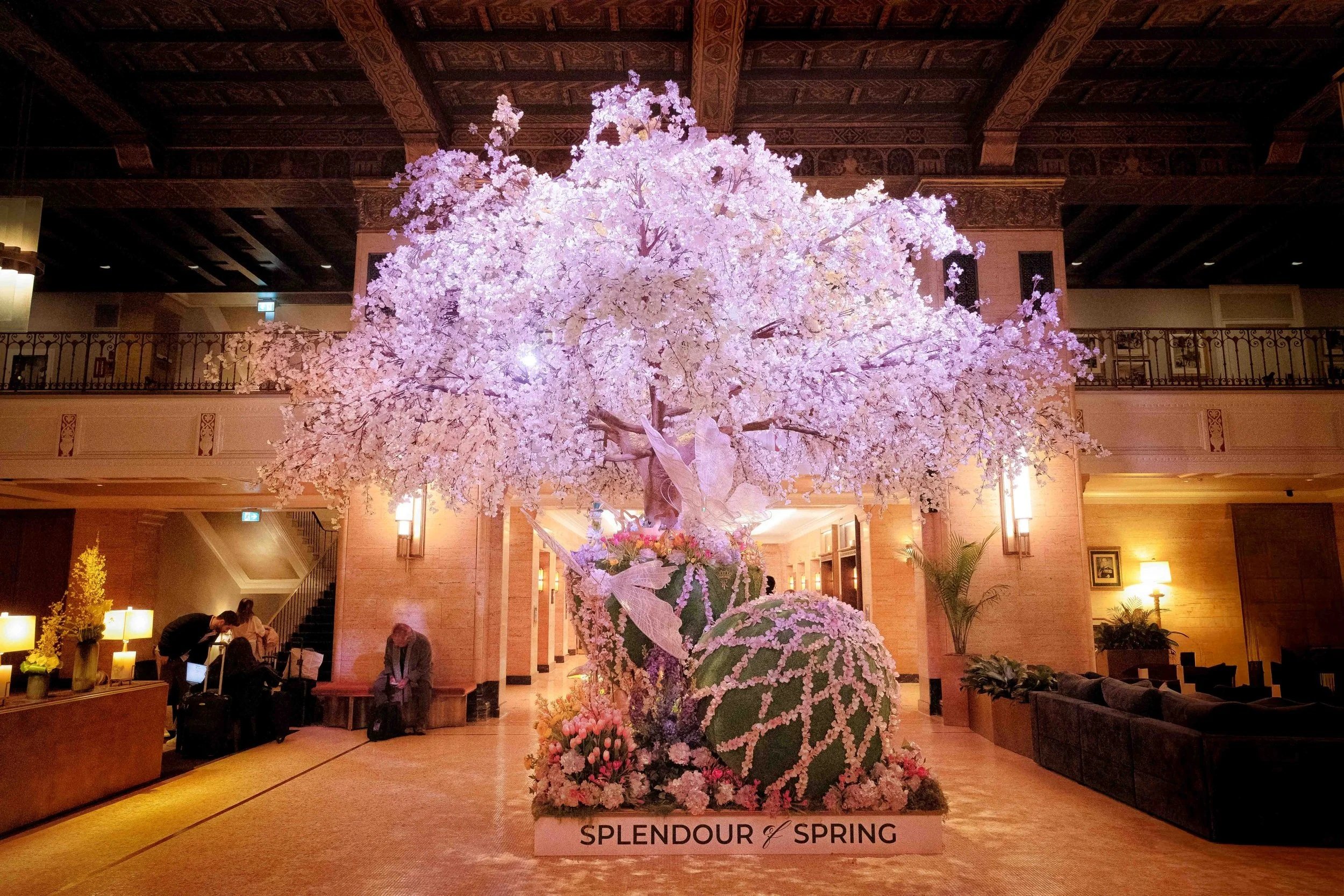 Front-facing view of the Splendour of Spring display at the Fairmont Royal York, where a sculptural cherry blossom tree rises from a base of spring florals, framed by the warm stone and architectural symmetry of the hotel lobby.