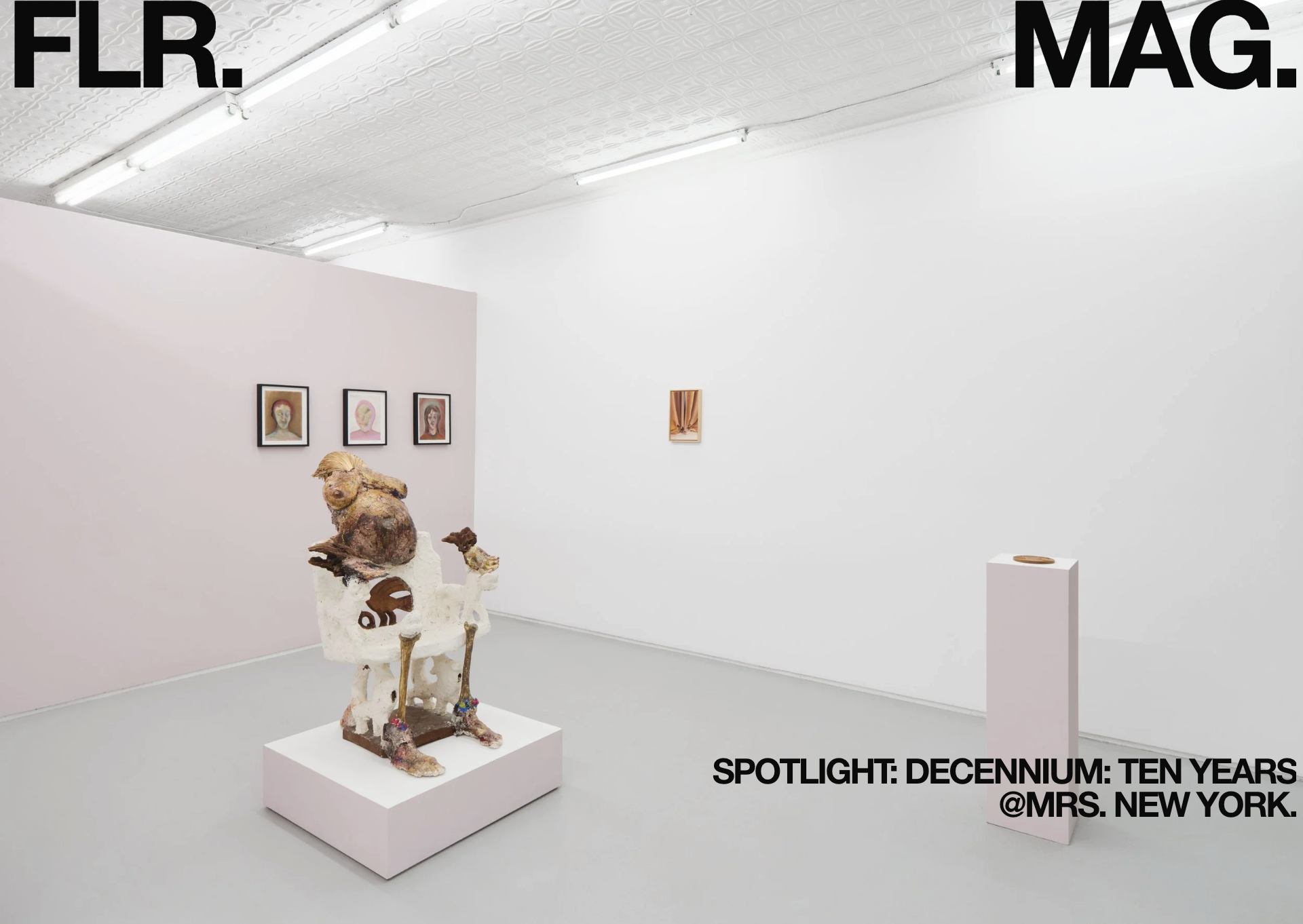 Art gallery exhibit featuring various artwork, including three framed portraits of people with colorful backgrounds on a light pink wall, a small framed piece of fabric, a sculpture resembling a seated animal with a person on its back, and a small pedestal with a circular art piece. The room has a white ceiling with fluorescent lights and plain white walls.