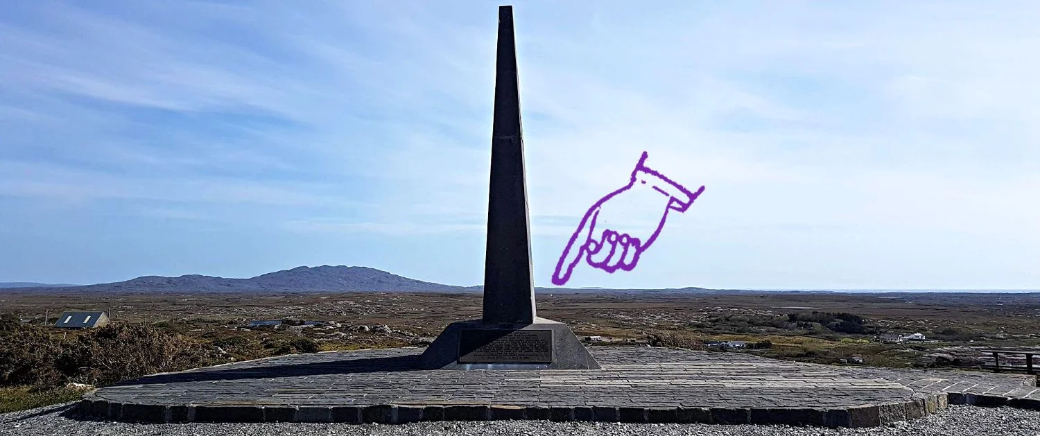 Alcock and Brown Memorial, end-on view. From the side it looks exactly like the tail-plane of a modern airliner. I’m regretting the pointing hand already but, depending on what you’re viewing this on, you should see a white dot on the moor below the finger. That’s the cairn marking the original crash site. The picture also gives an idea of the scale of the Derrigimlagh Bog as well.
