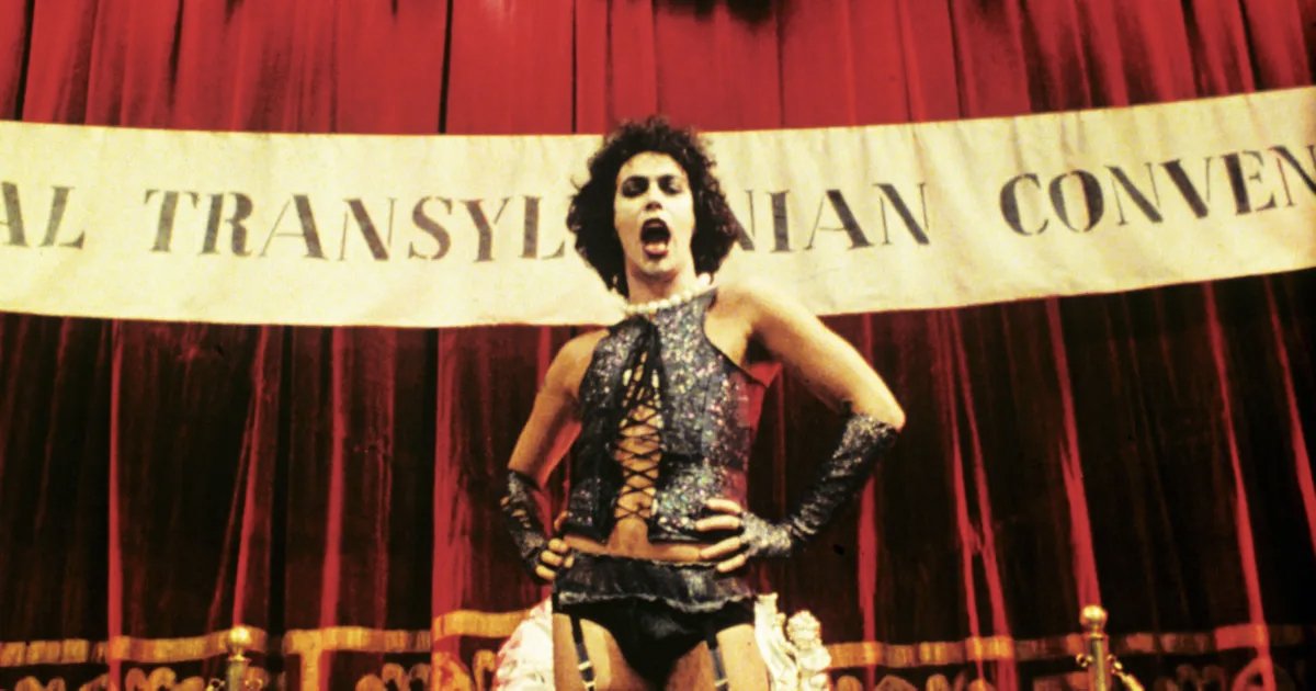 A white man in lingerie with his hands on his hips in front of a red curtain with a banner.