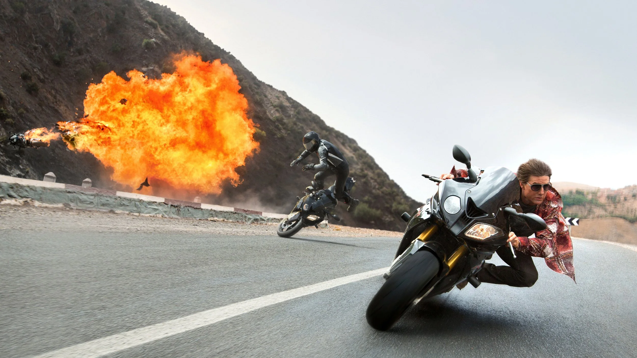 A dark-haired man on a motorcycle leaning at a 45 degree angle to the highway. Behind him, an explosion and a man flying from his motorcycle.