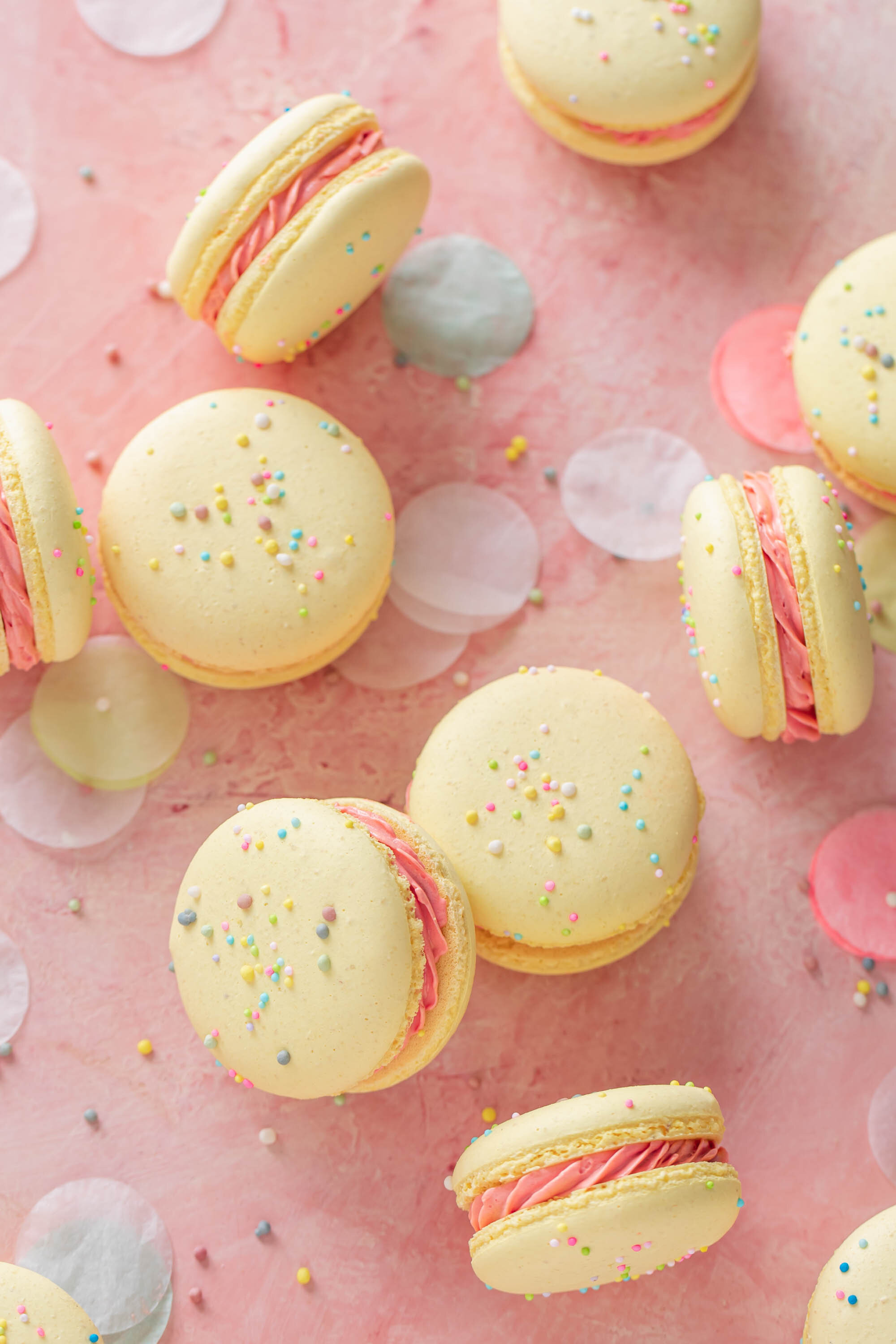 Pastel French macarons with sprinkles and pink buttercream filling scattered on a pink table with confetti.