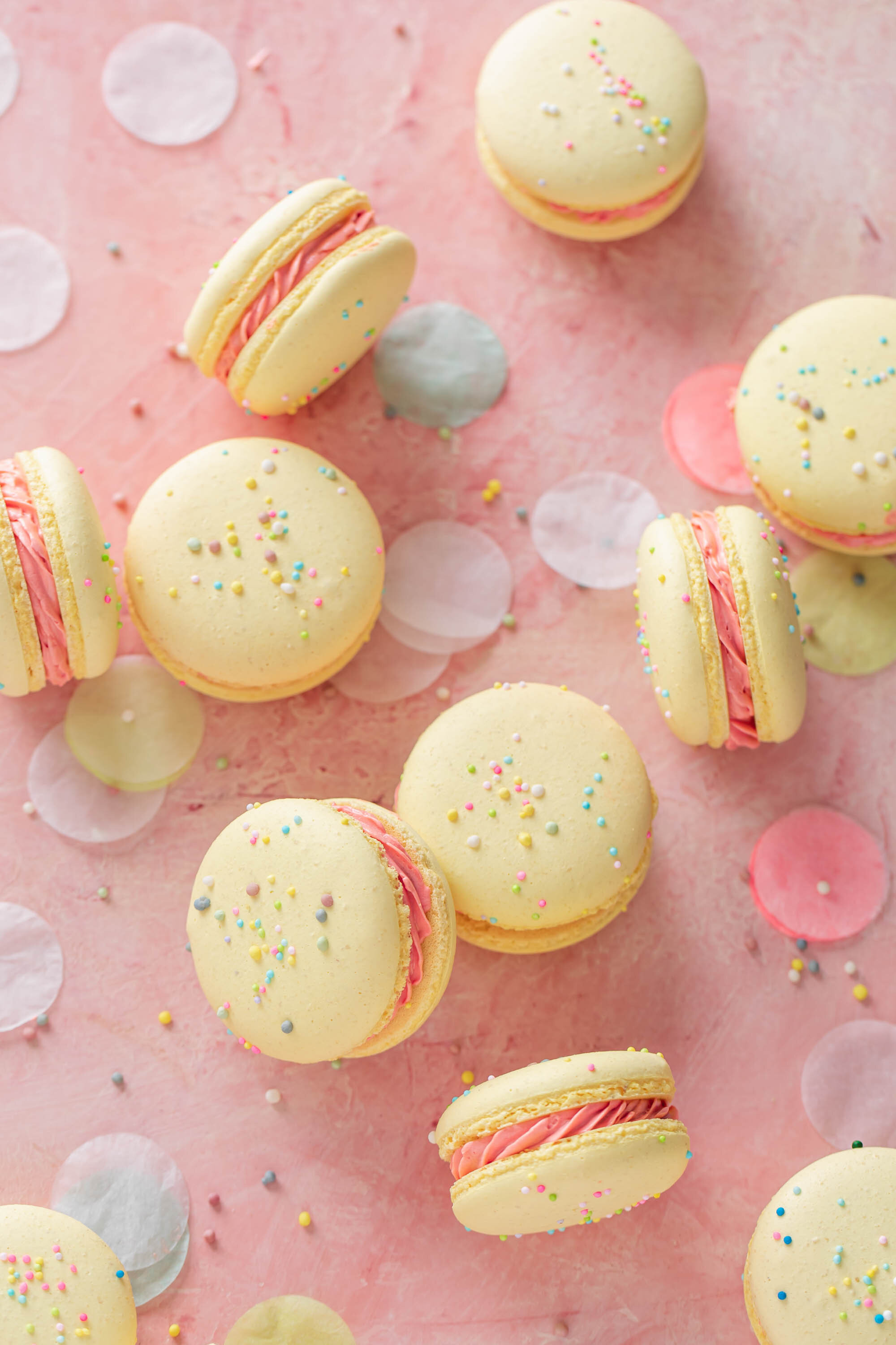 French macarons with sprinkle and pink buttercream filling scattered on a pink table with confetti.