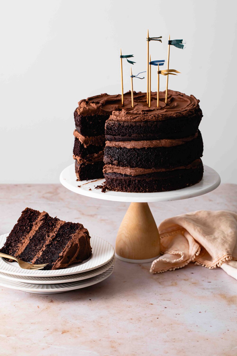 An 8-inch chocolate layer cake with fluffy fudge frosting set on a white and wood cake stand with one slice removed.
