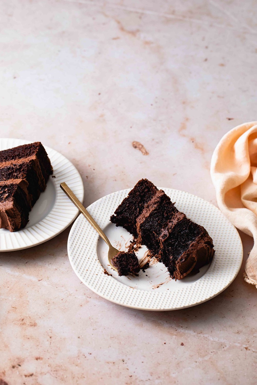 A half eaten slice of moist chocolate cake with fluffy fudge frosted set on a creamy marble tabletop.