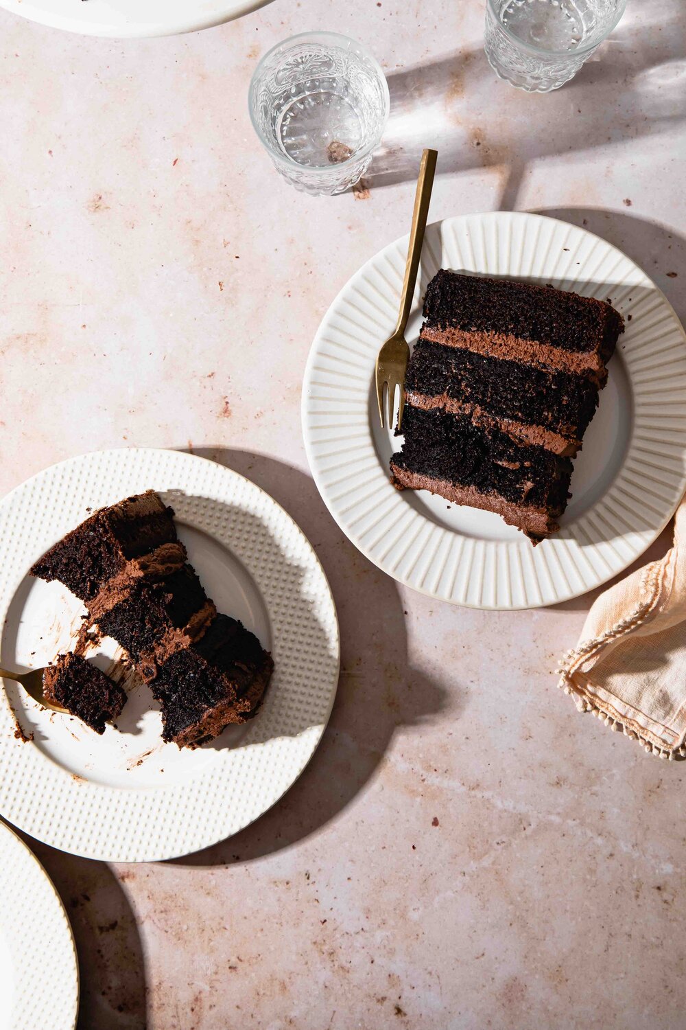 A photo of slices of moist chocolate cake with fudge frosting wtih sunlight and harsh shadows set on cream colored marble table.