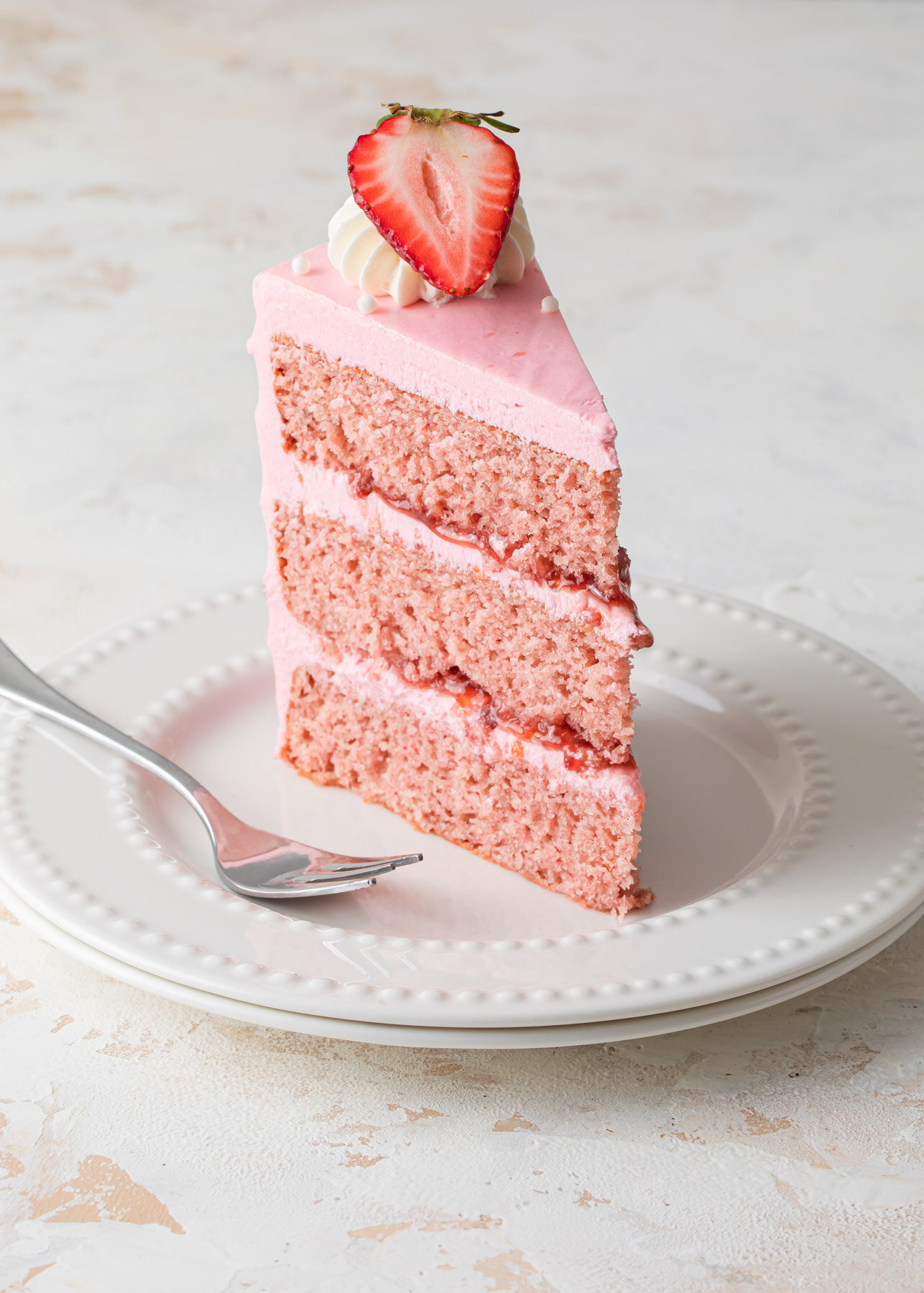 A slice of  Strawberry Layer Cake with strawberry jam and pink ermine frosting on a white plate.