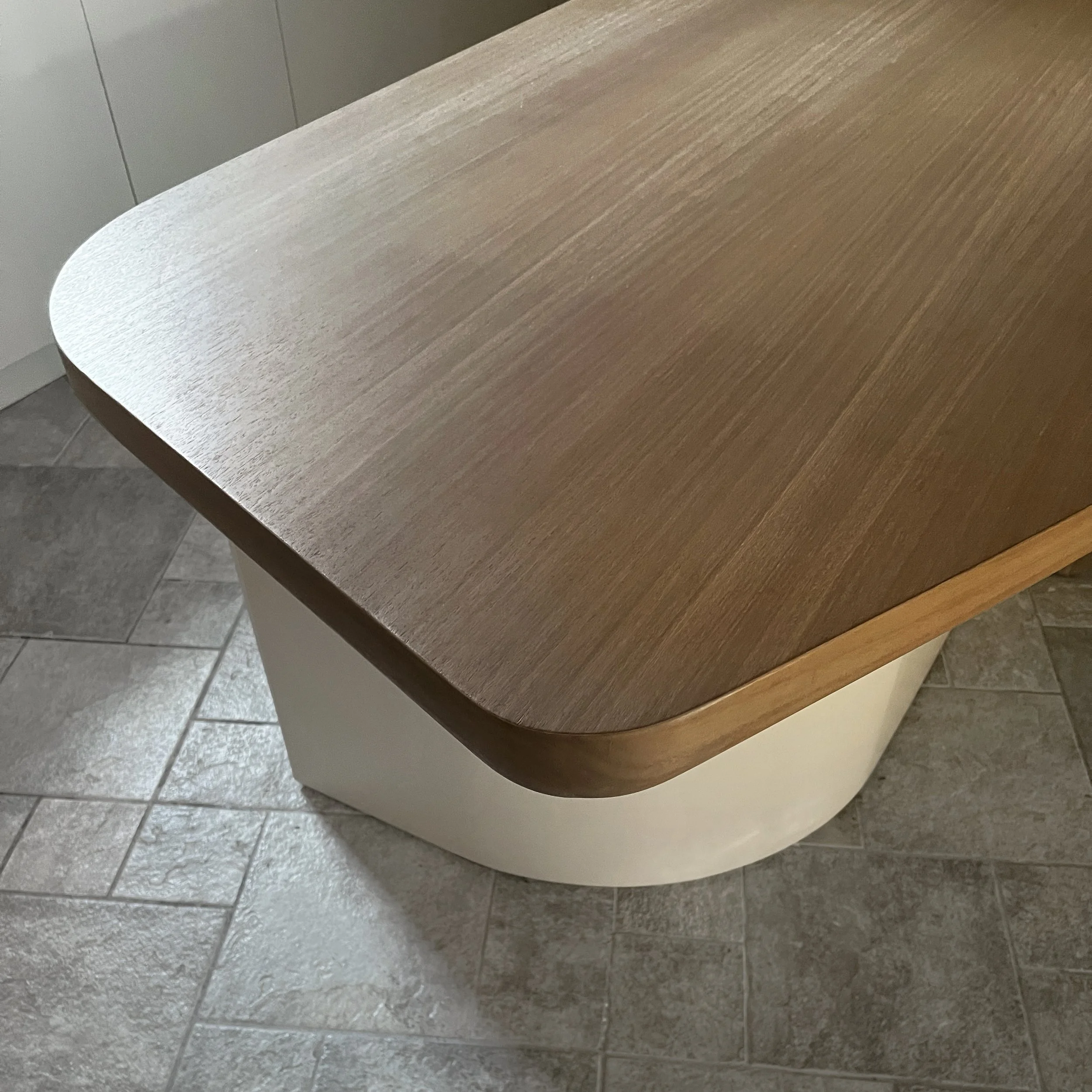 Close-up of a wooden-topped table with a rounded edge, supported by a white, rounded base, on a tiled floor.