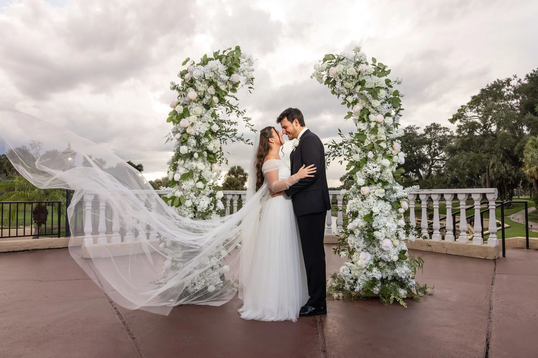 Rachel and Paul embracing between white floral pillars at Mission Resort & Club, with Rachel’s veil flowing to the left.