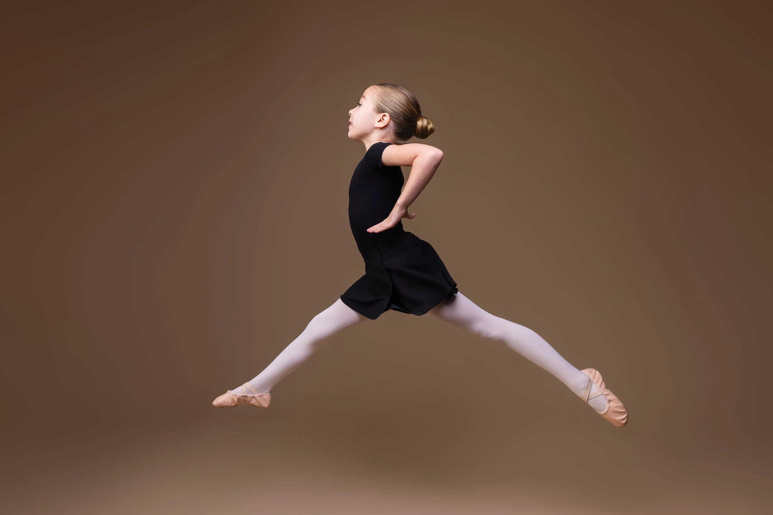 Young ballet dancer performing a dramatic leap against a warm brown studio backdrop, photographed by Dreamscape Photography + Video.