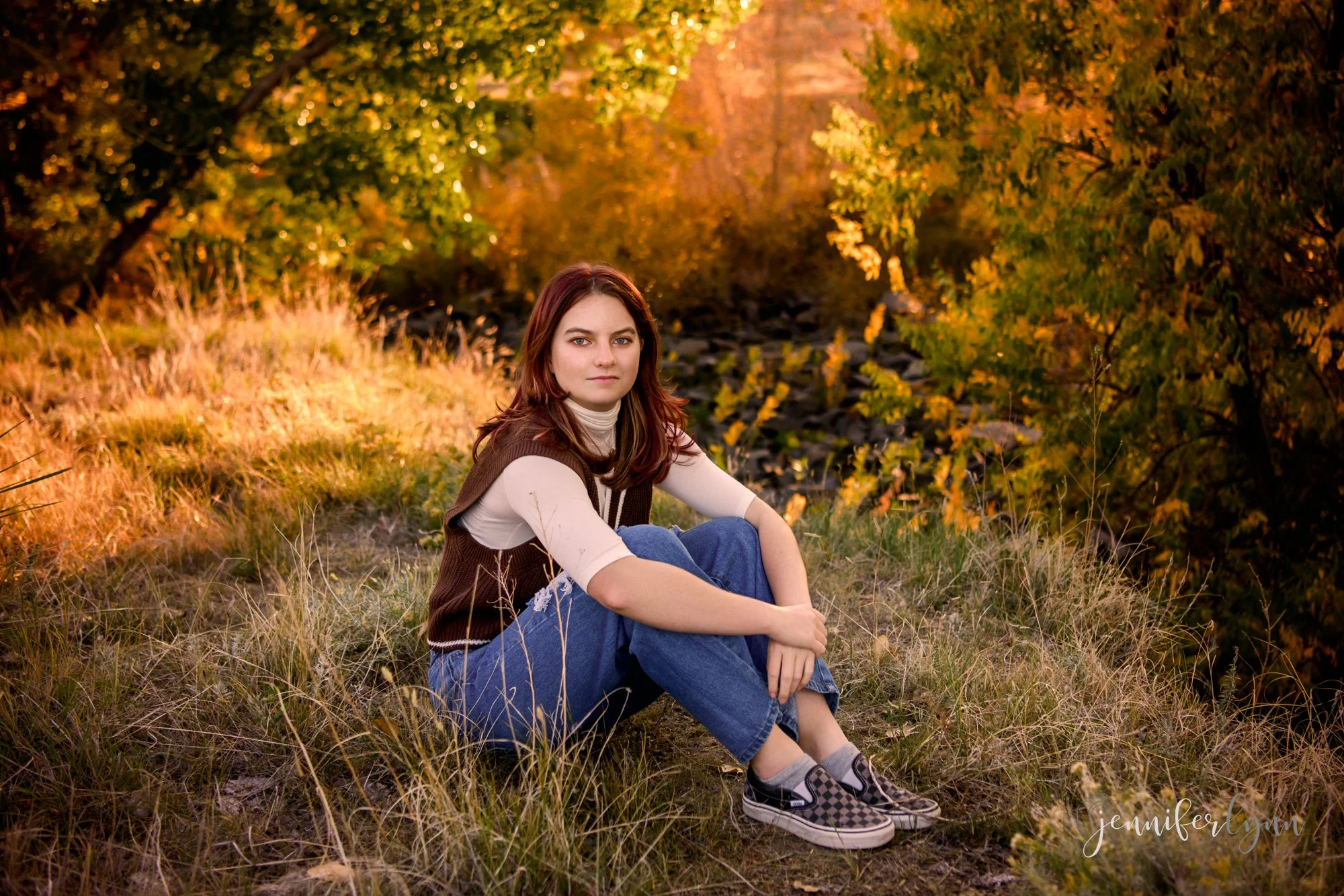 High school senior girl sitting in natural grass area with fall colors and golden light behind her